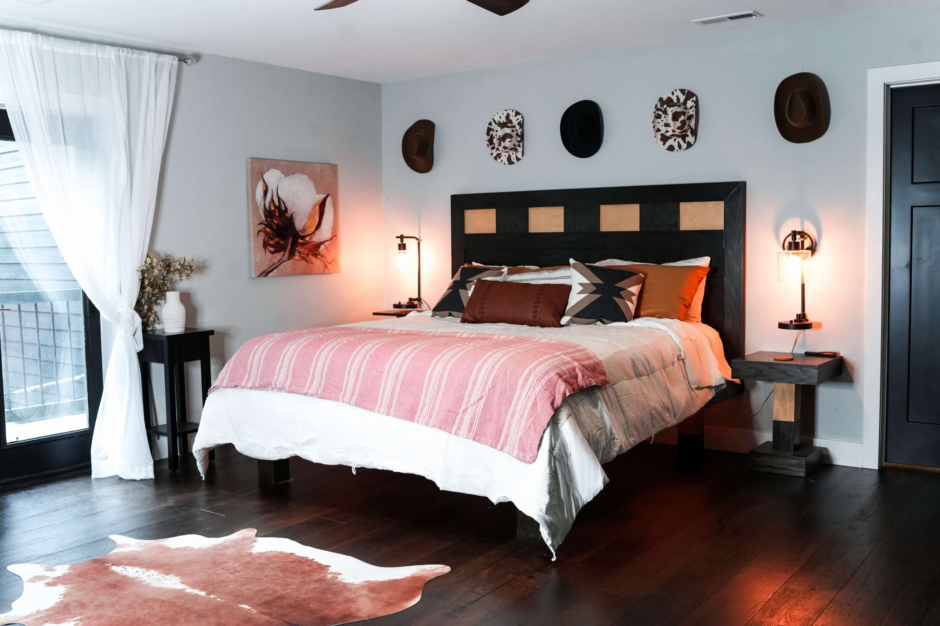 The image shows a modern bedroom featuring a large bed with patterned bedding, a cowhide rug on dark wooden flooring, and wall decorations including hats and artwork.