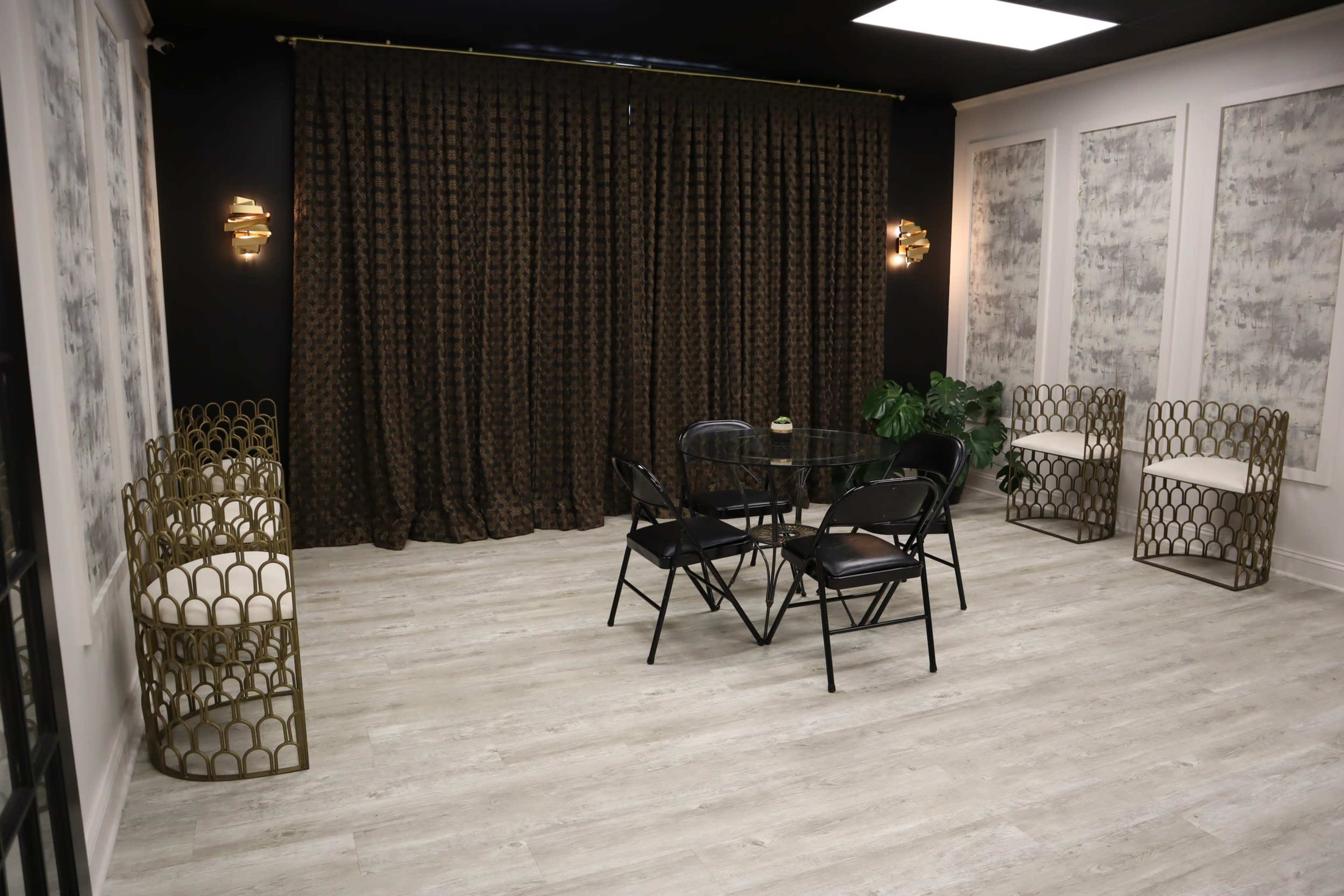 The image shows a modern interior space featuring a round glass table with black chairs, decorative gold seating, and dark curtains against patterned walls.