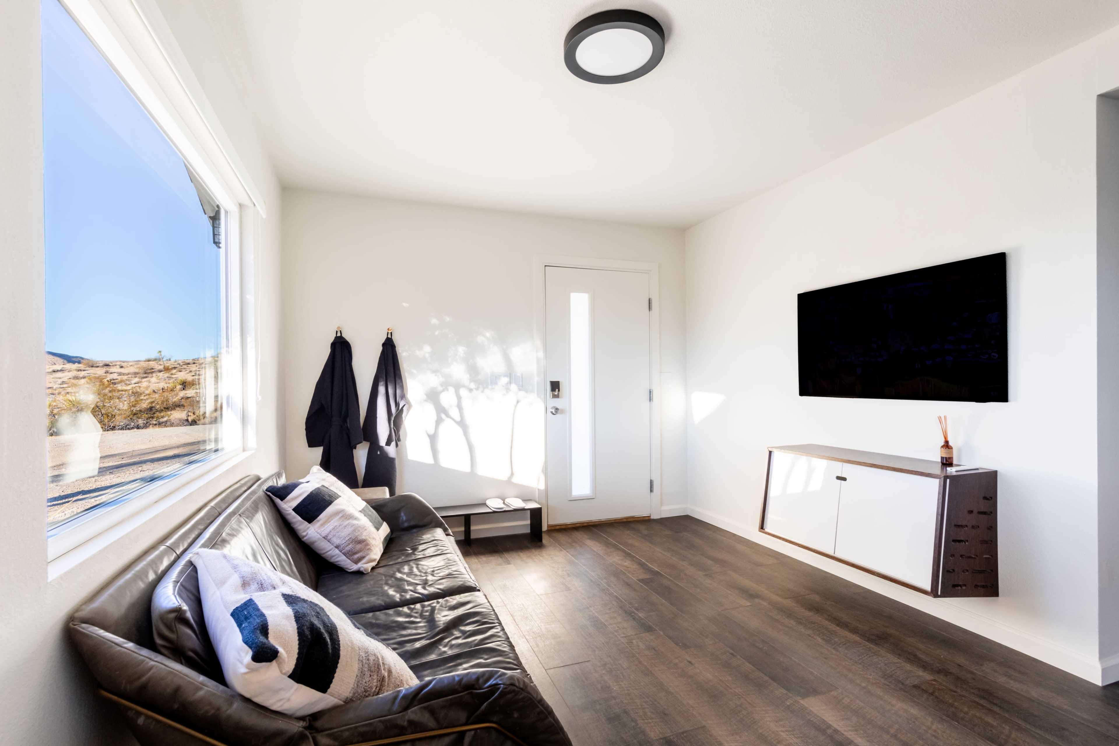 A modern living area features a black leather sofa, a wall-mounted television, and a white cabinet, with natural light coming through a large window.