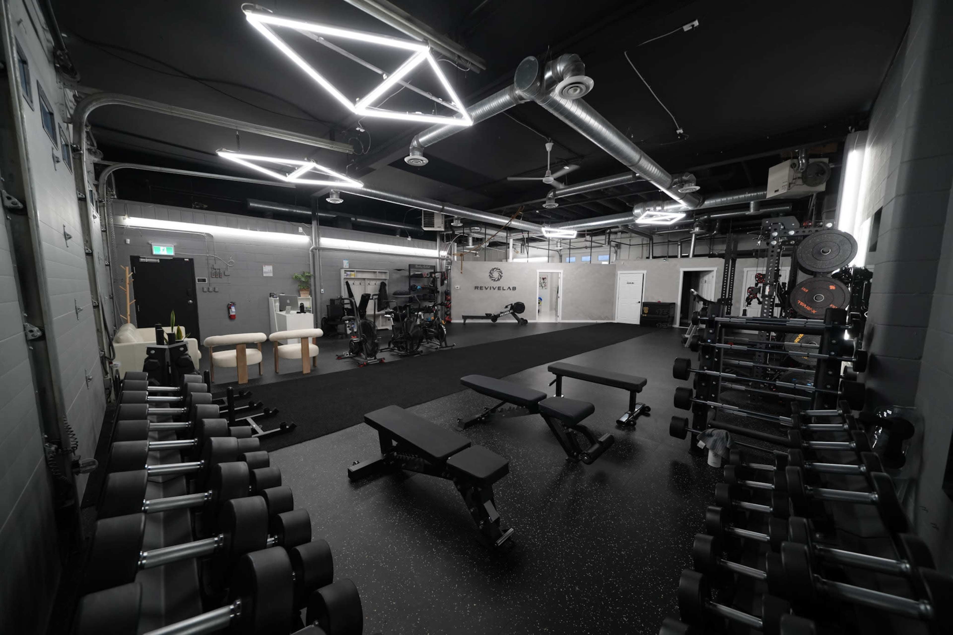 The image shows a well-organized gym space featuring weight benches, dumbbells, and various exercise equipment under modern lighting.