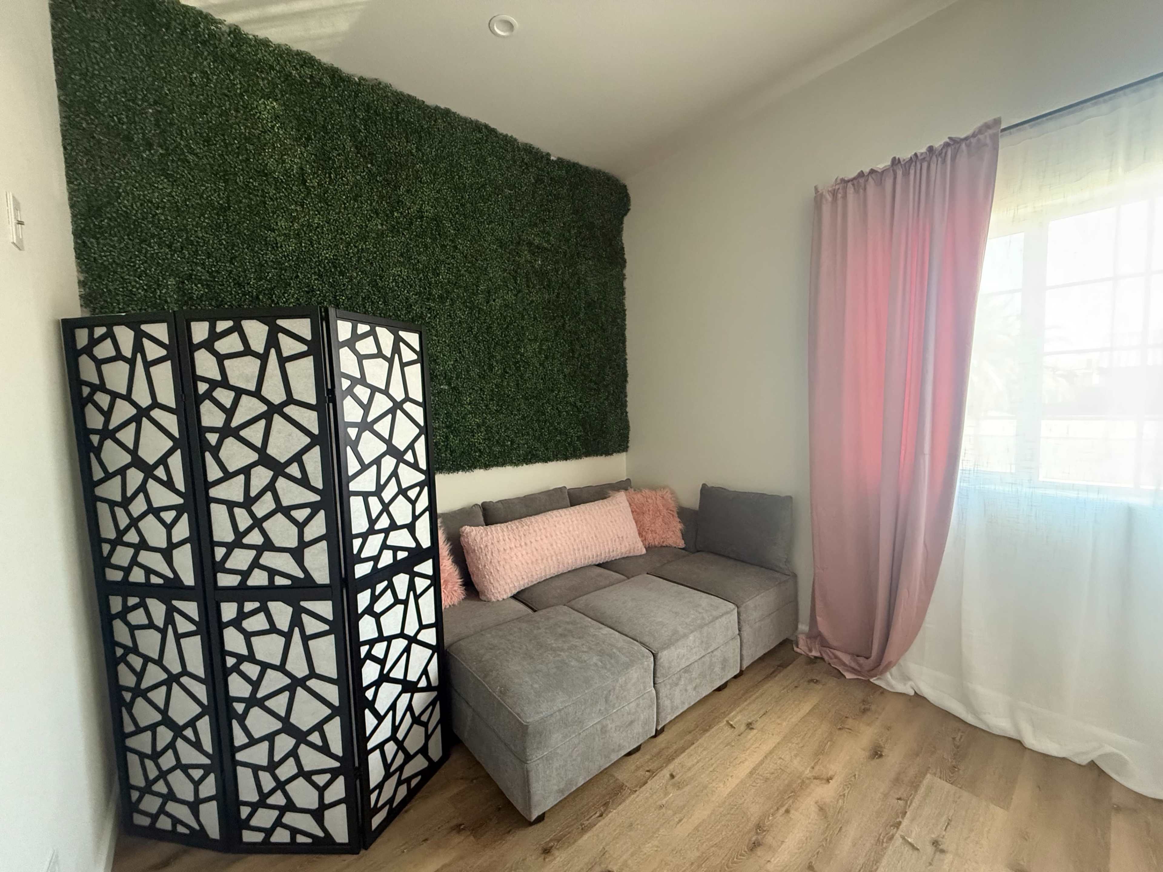 The image features a cozy living area with a gray sectional sofa, a decorative screen, and a green wall accent, complemented by light-colored curtains and wooden flooring.