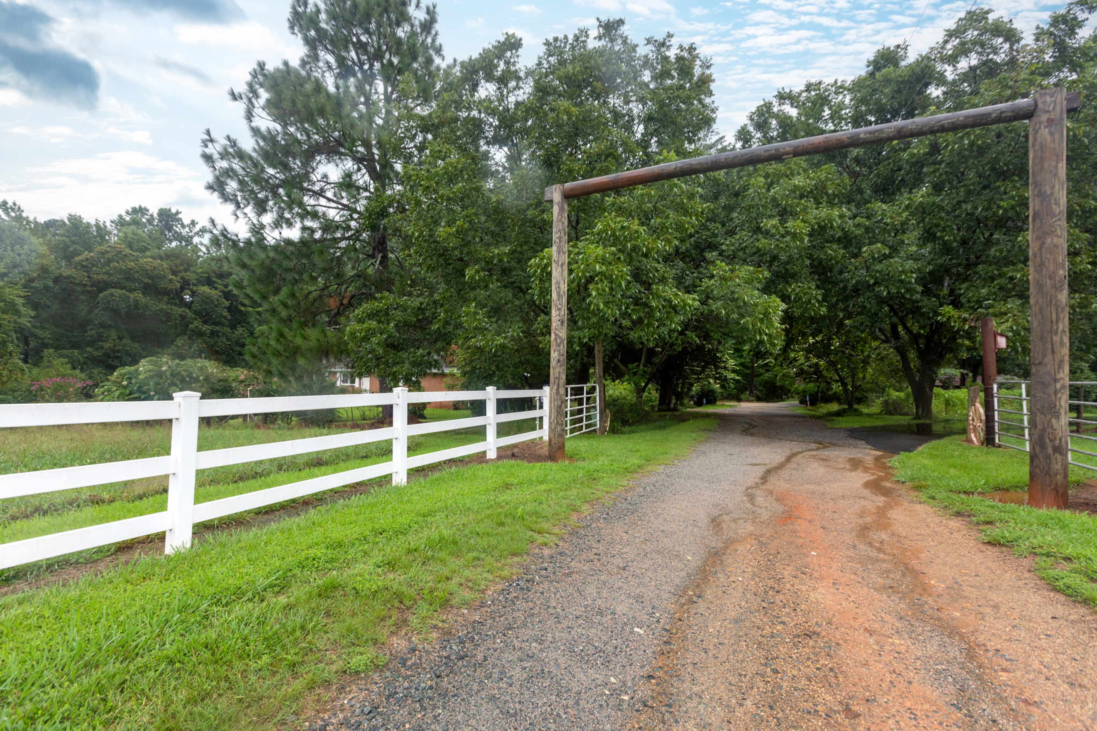 A gravel road leads through a wooden entrance arch, framed by lush greenery and a white fence on either side.