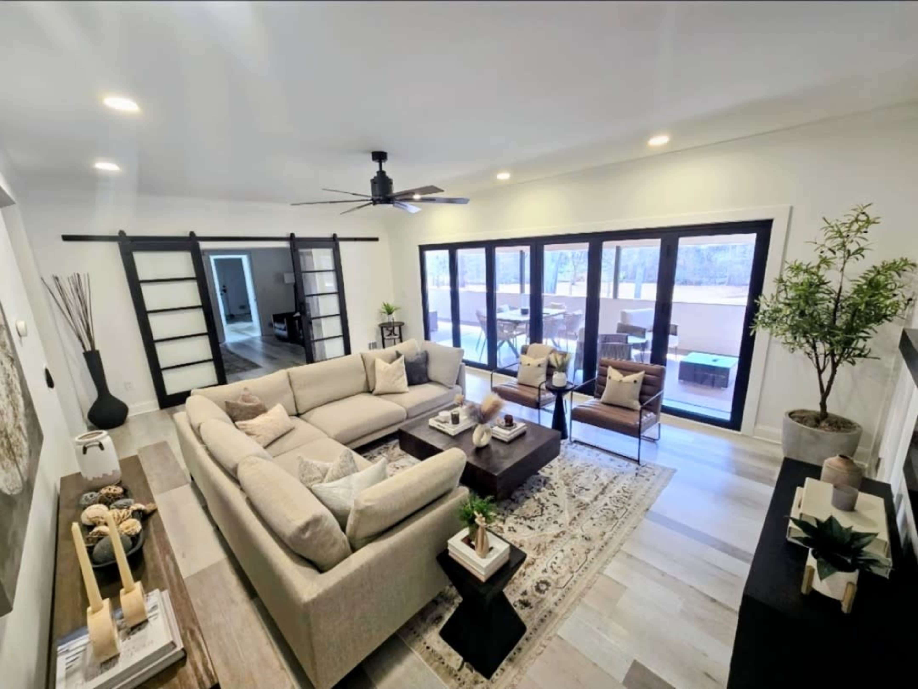 The image shows a modern living room featuring a large sectional sofa, a coffee table, and sliding glass doors that open to an outdoor area, with decorative plants and a stylish rug.