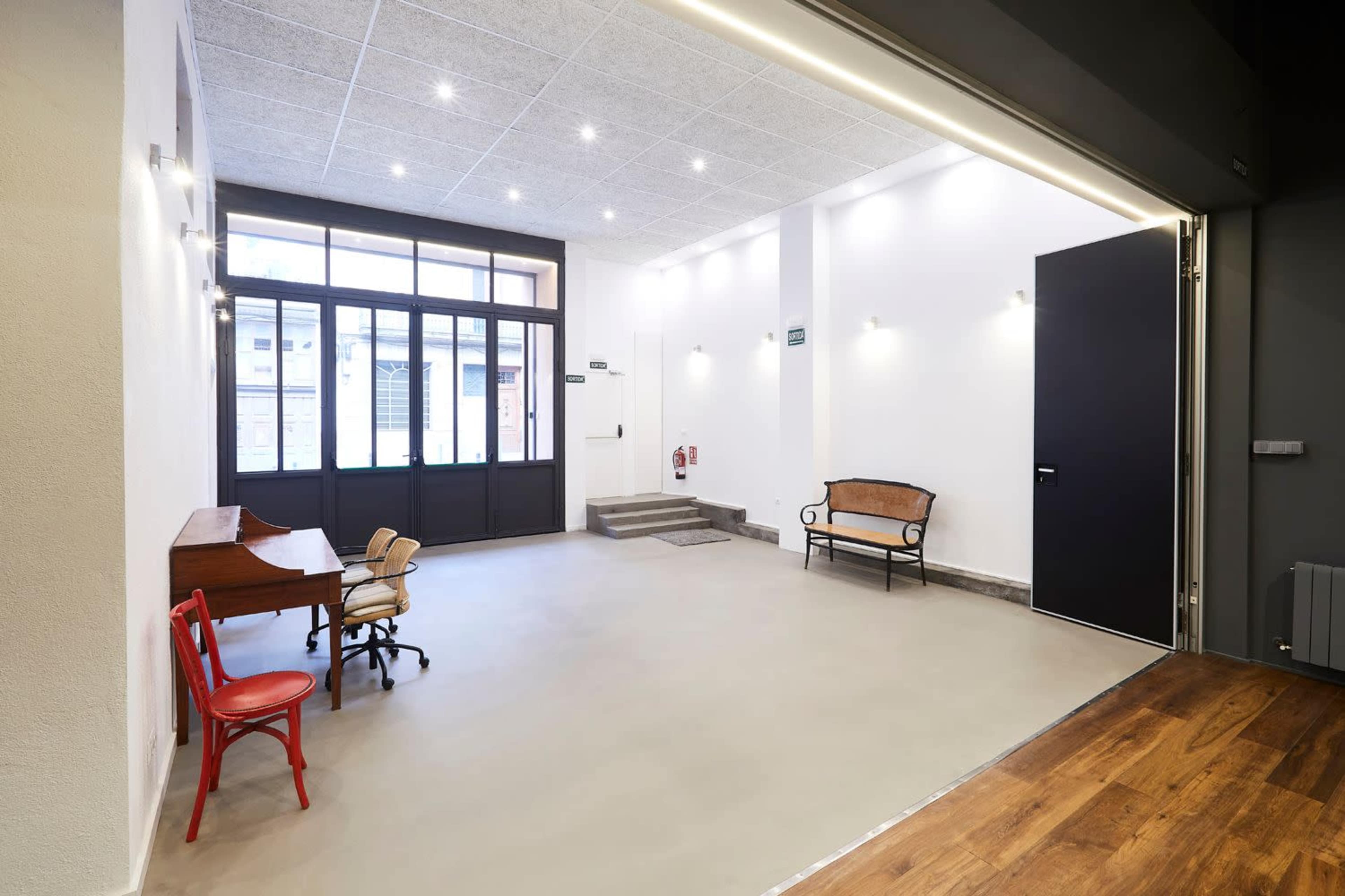A spacious lobby area with a wooden bench, two chairs, and a staircase, featuring large windows that allow natural light to enter.