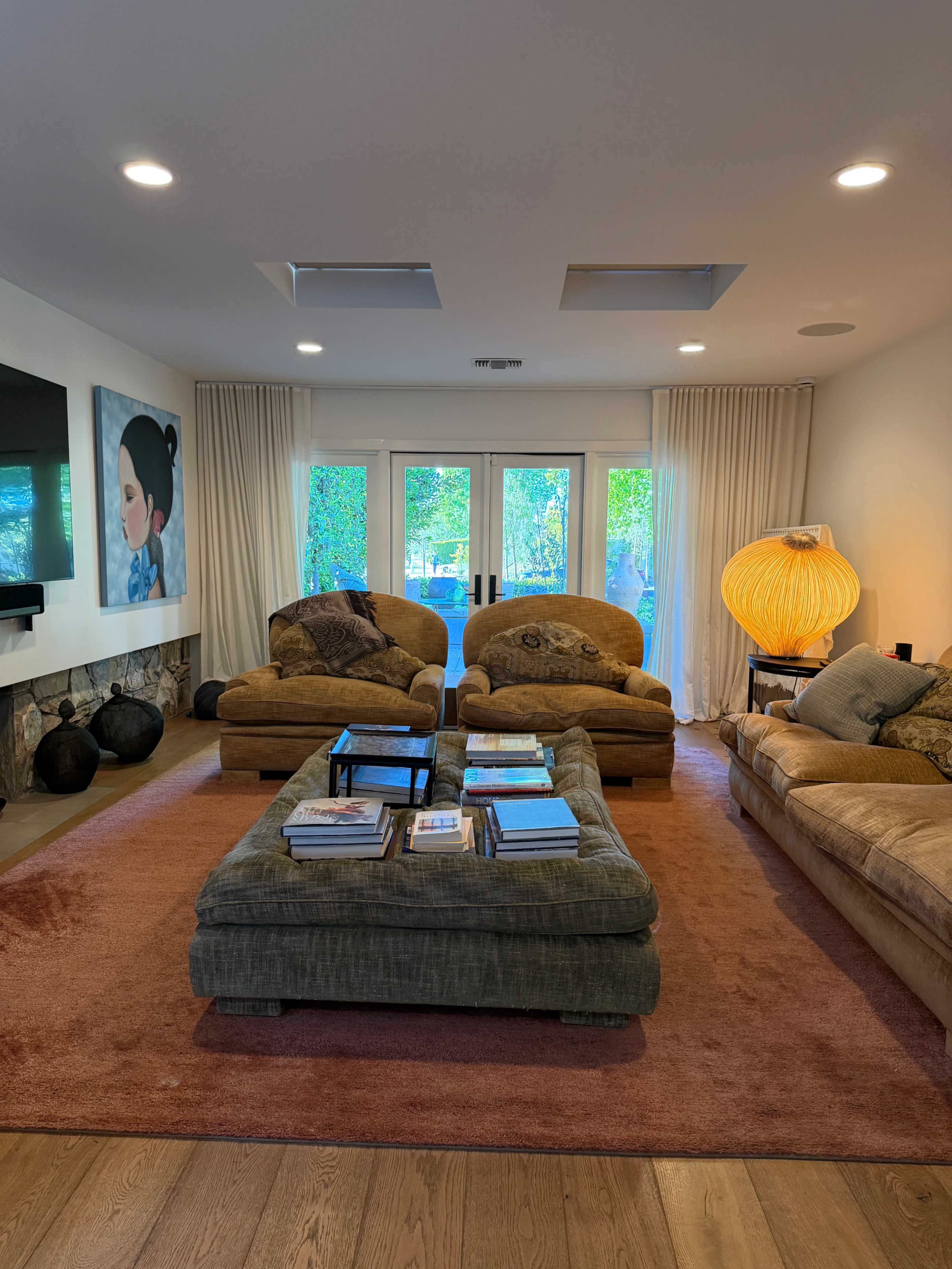 The image shows a spacious living room with two large sofas, a coffee table stacked with magazines, a flat-screen TV, and large windows overlooking a green outdoor area.