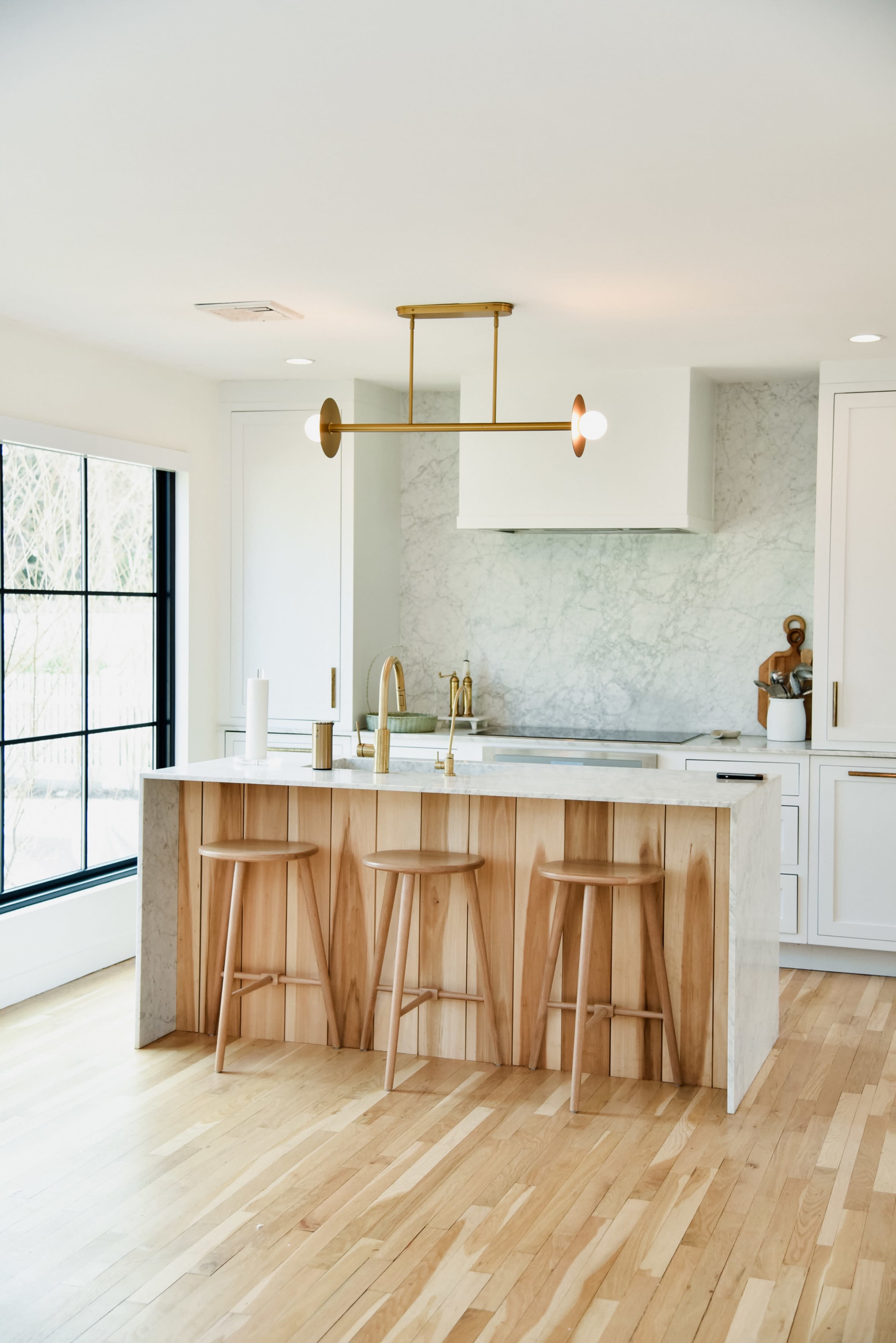 The image shows a modern kitchen featuring a marble backsplash, a central island with wooden accents, and three bar stools.