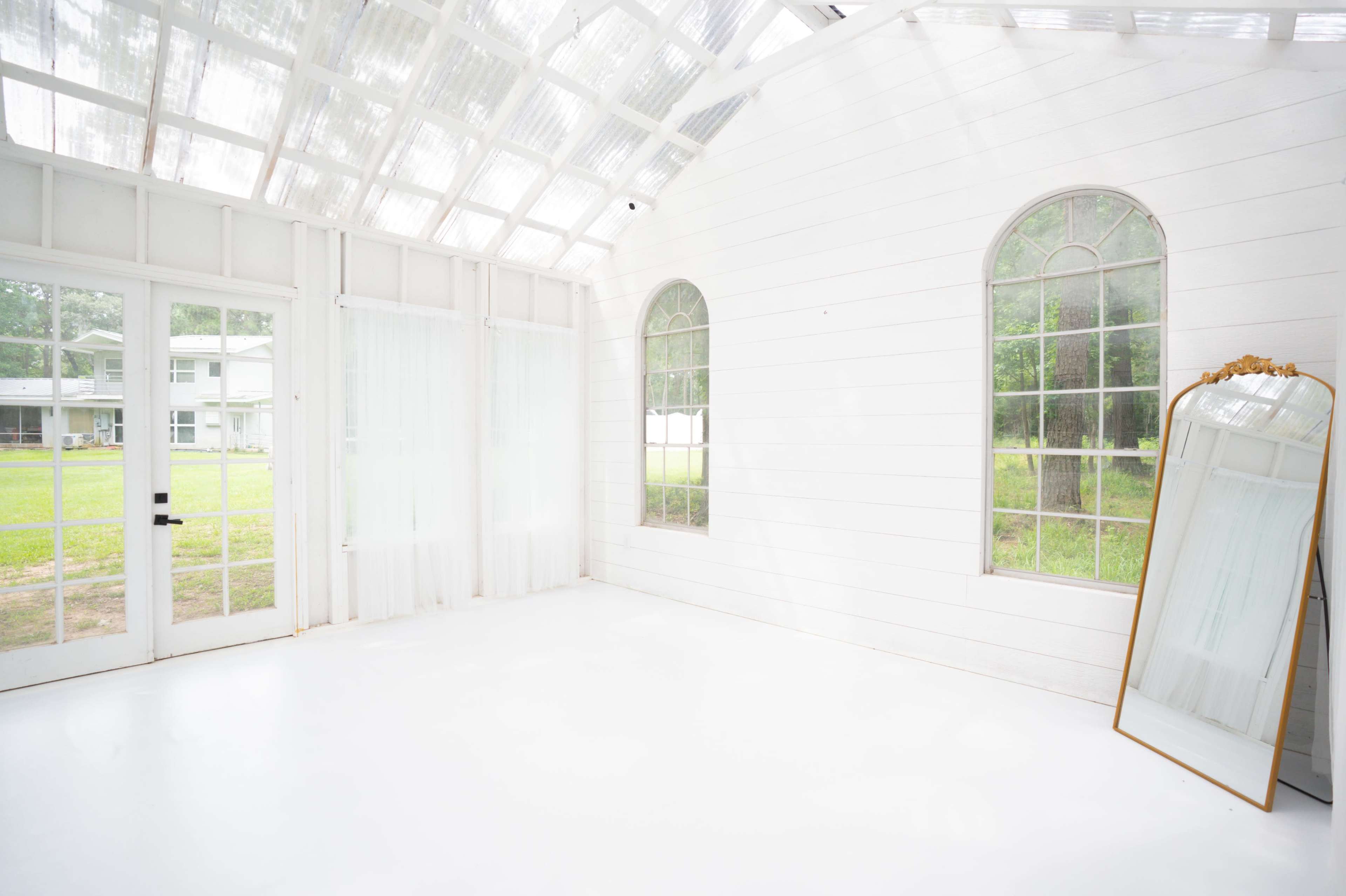 A bright, open room features clear windows, white walls, and a large mirror, with a view of an outdoor grassy area through the glass doors.