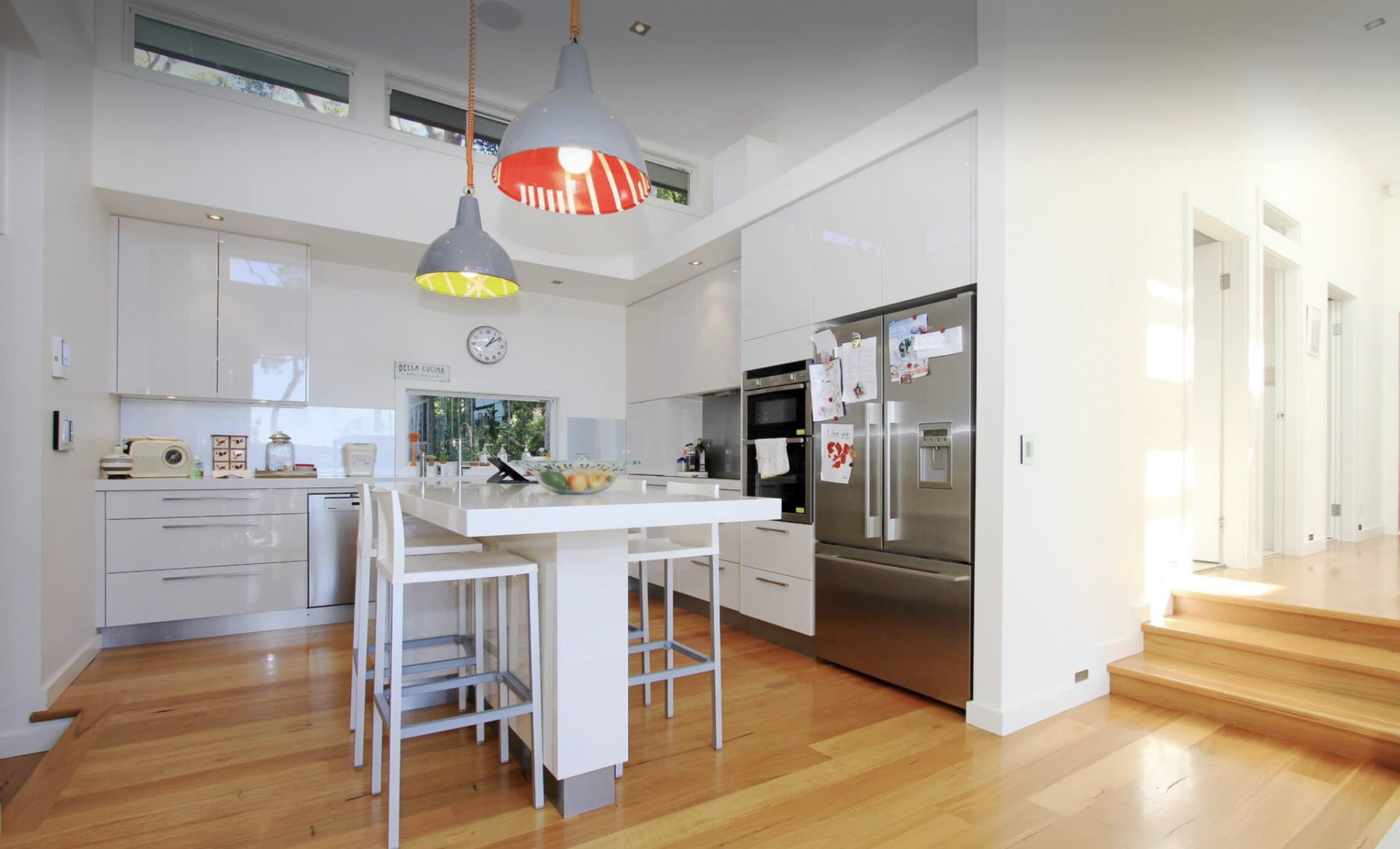 The image shows a modern kitchen with white cabinetry, a central island with bar stools, stainless steel appliances, and wooden flooring.