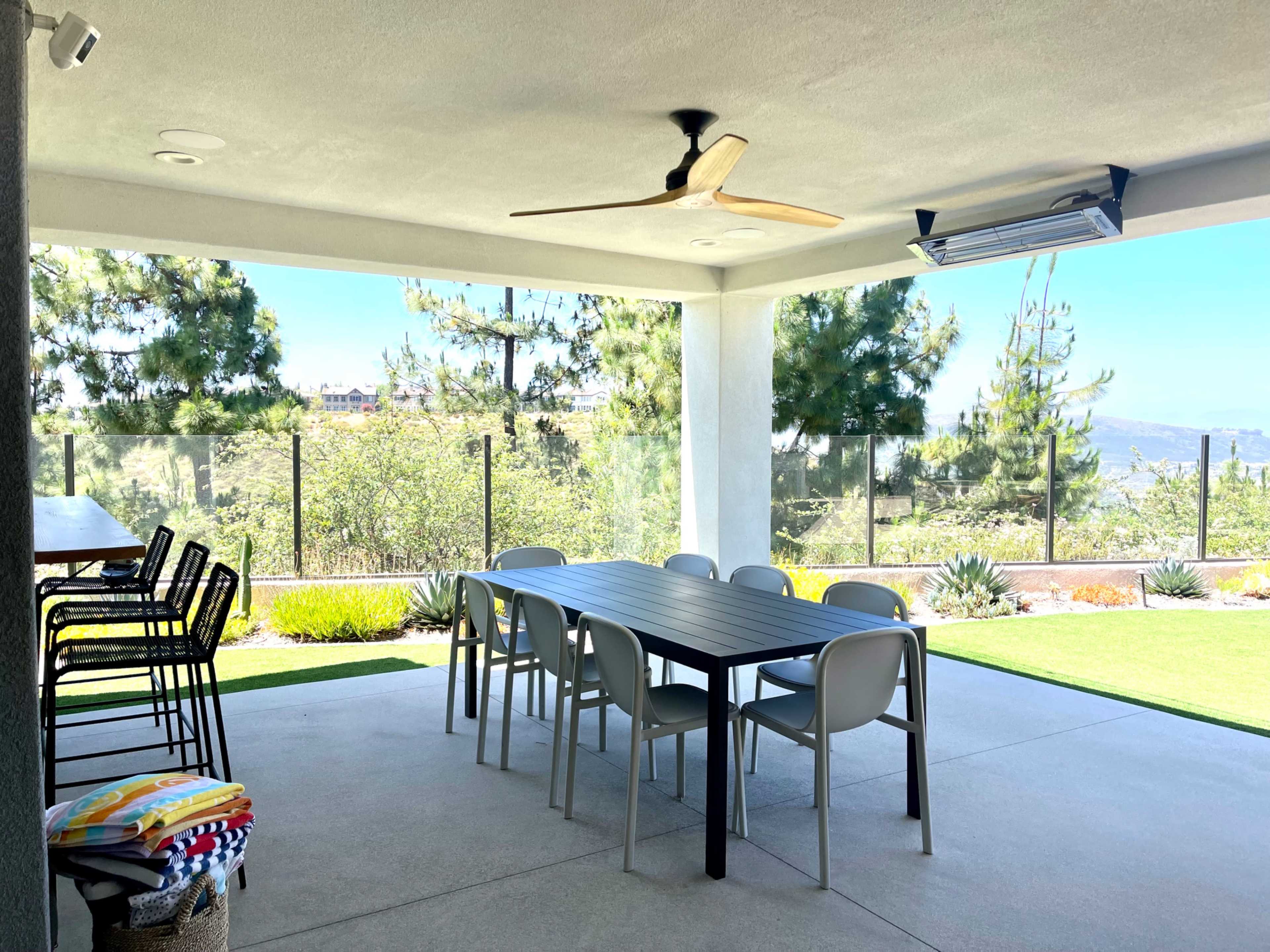 A covered patio features a long dining table with chairs, surrounded by greenery and a view of distant hills.