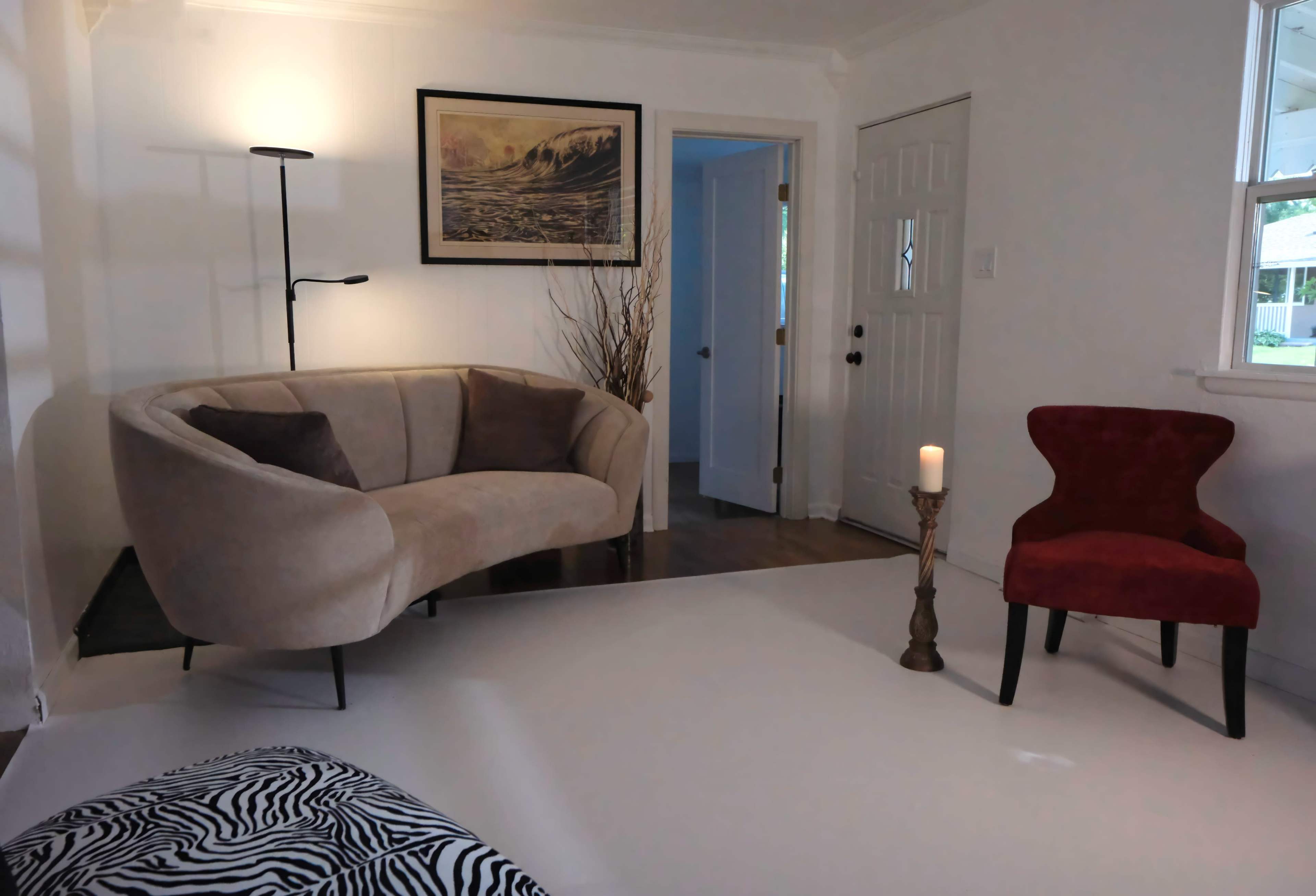 The image features a cozy living room with a beige curved sofa, a red accent chair, a candle on a stand, and a large framed landscape picture on the wall.