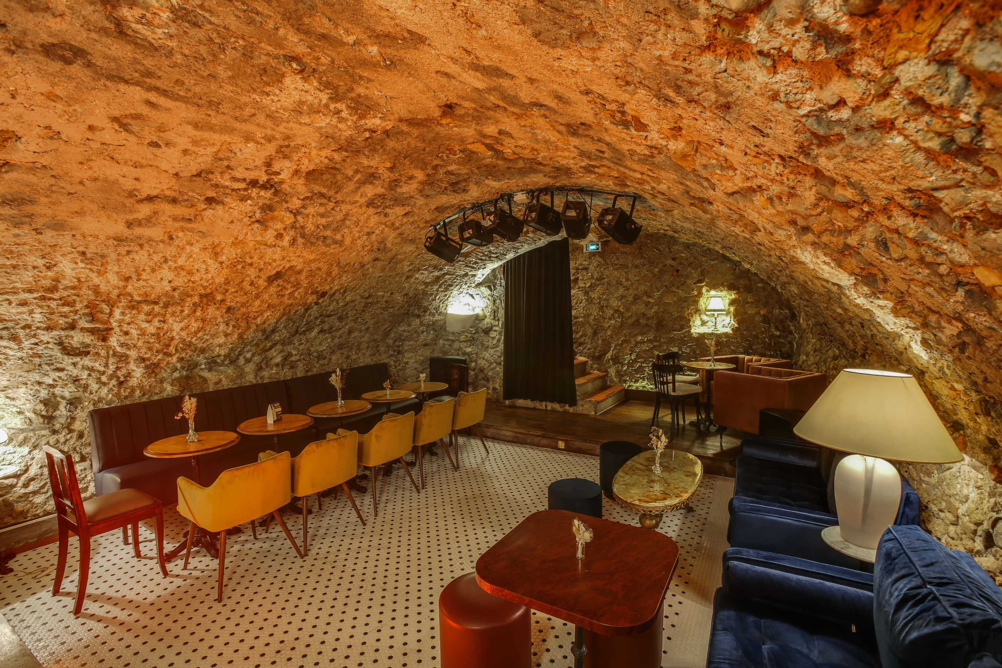 The image shows a cozy underground bar with stone walls, featuring seating areas with tables and a stage illuminated by spotlights.