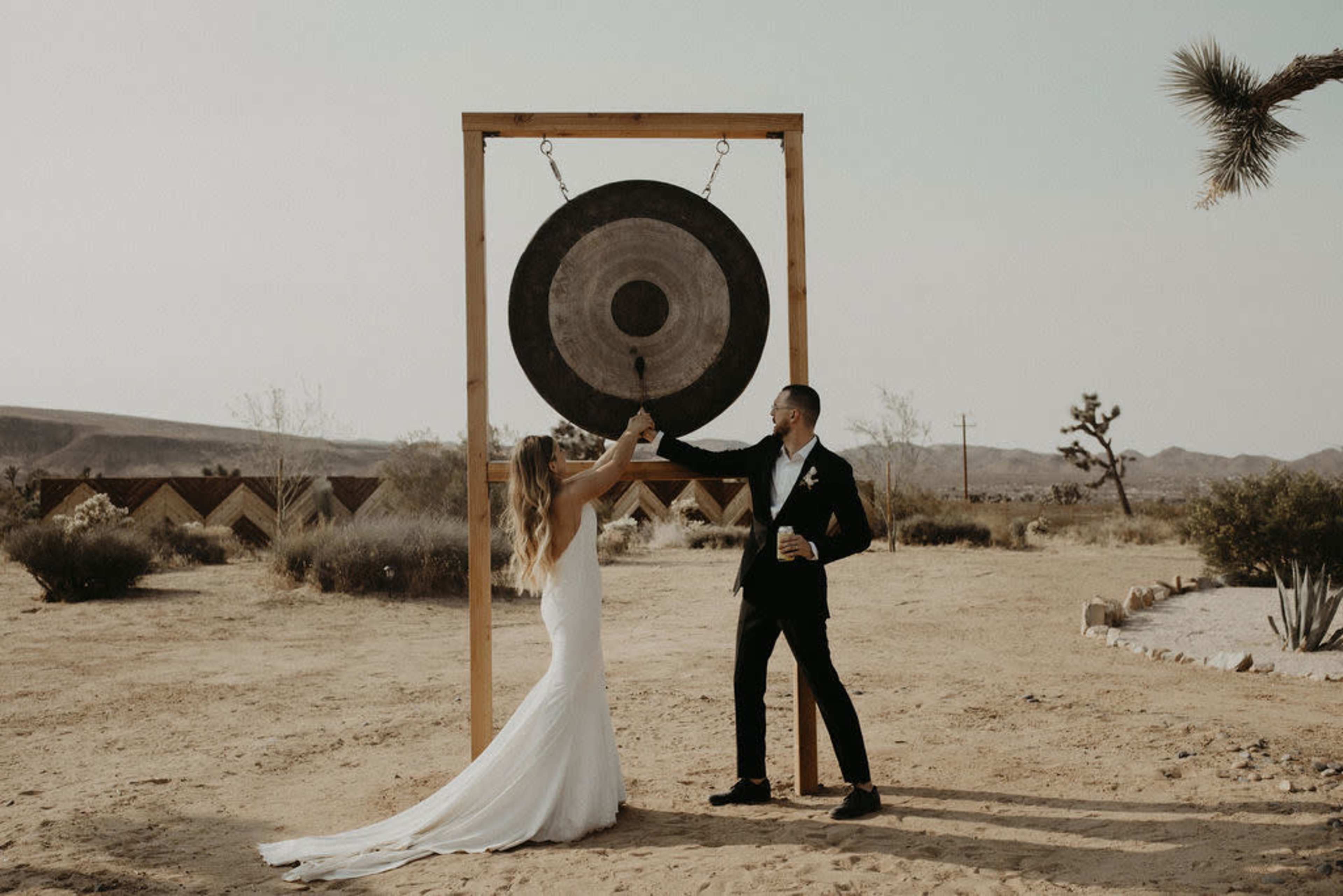 A couple in formal attire strikes a large gong in a desert setting, with desert plants and mountains in the background.