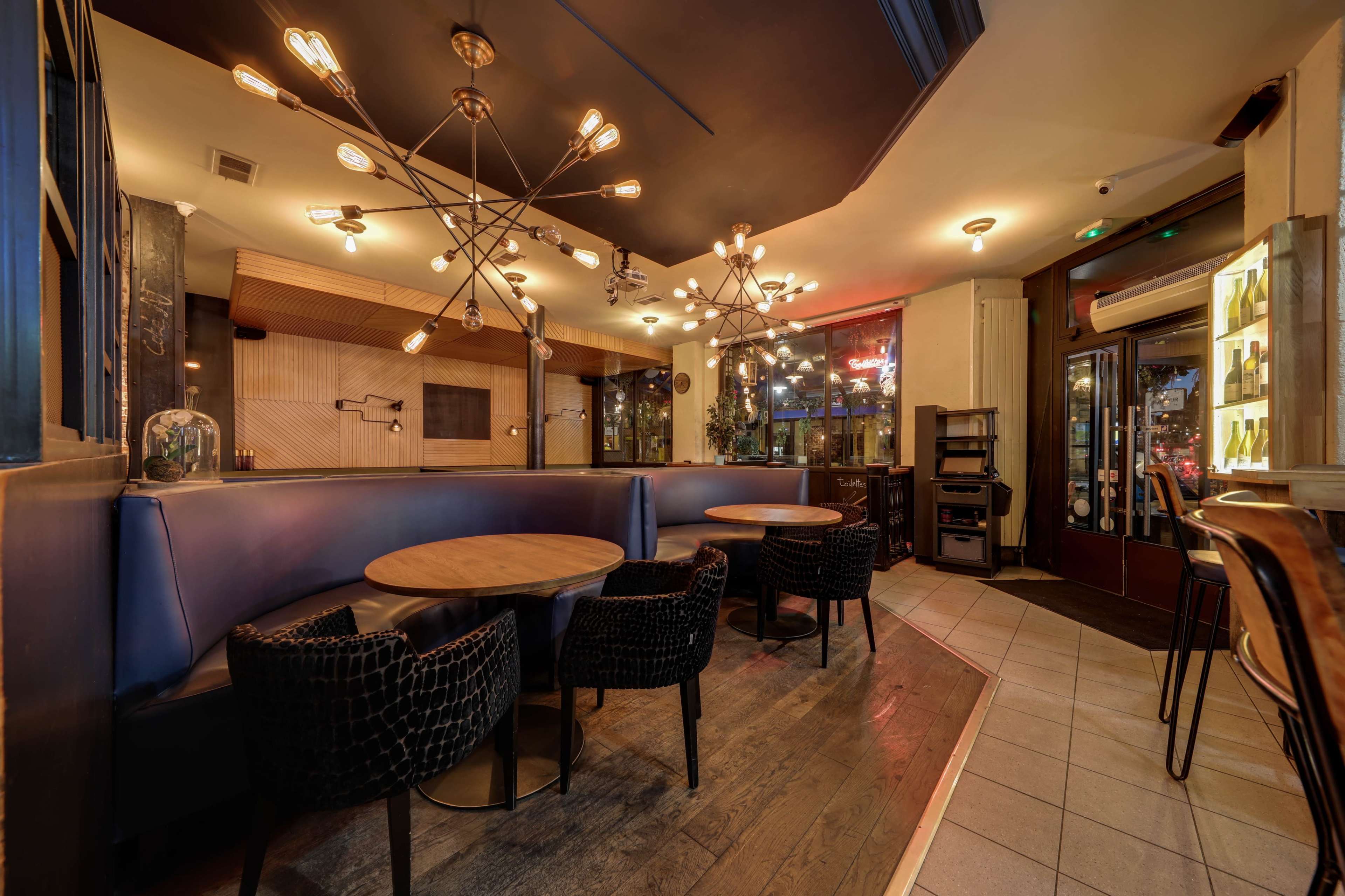 The image shows a modern restaurant interior with round wooden tables, black woven chairs, and statement lighting fixtures hanging from the ceiling.