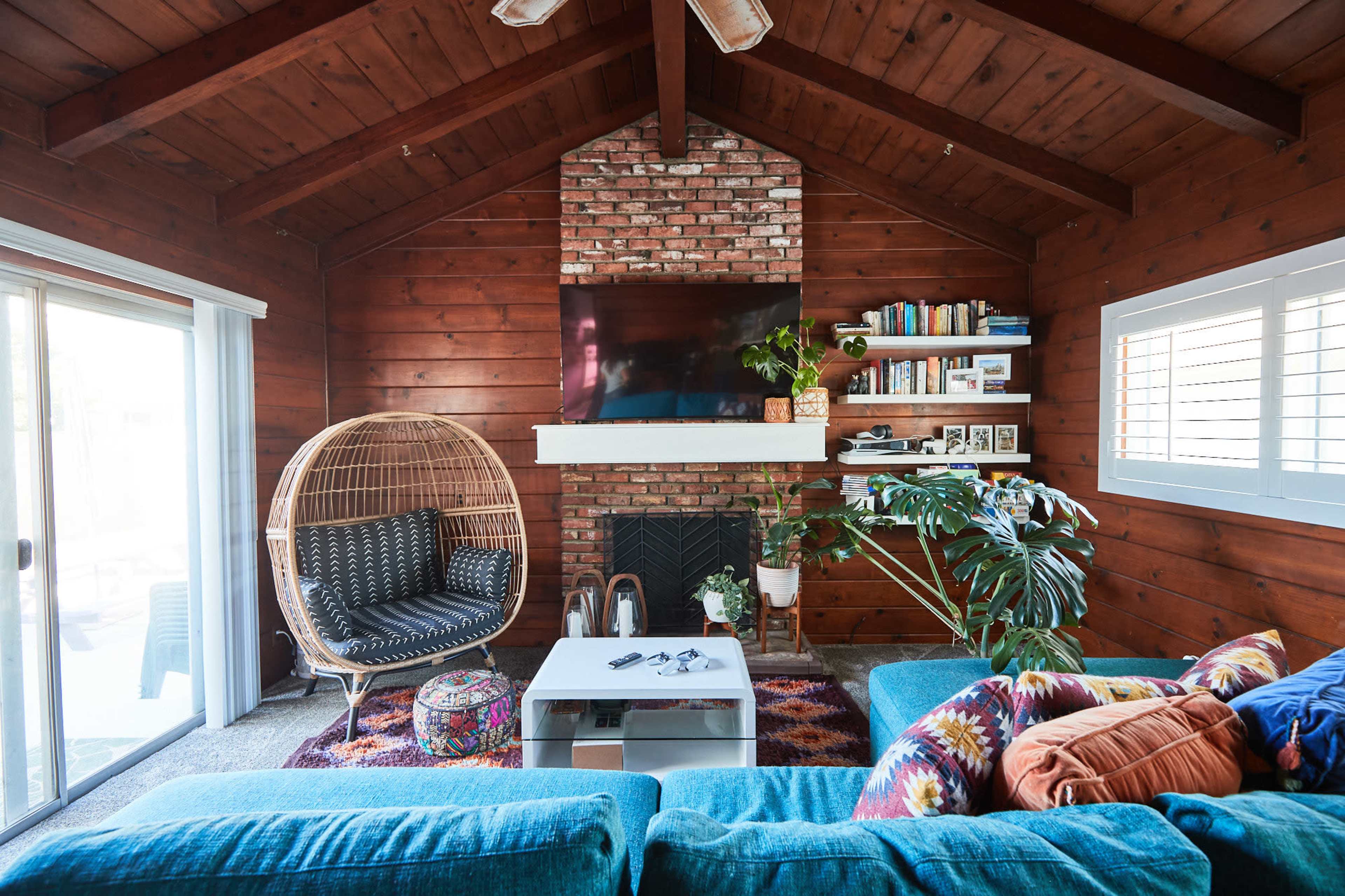 A cozy living room with wooden walls, a brick fireplace, a large television mounted above the mantel, and colorful furniture, including a blue sofa and a round chair.
