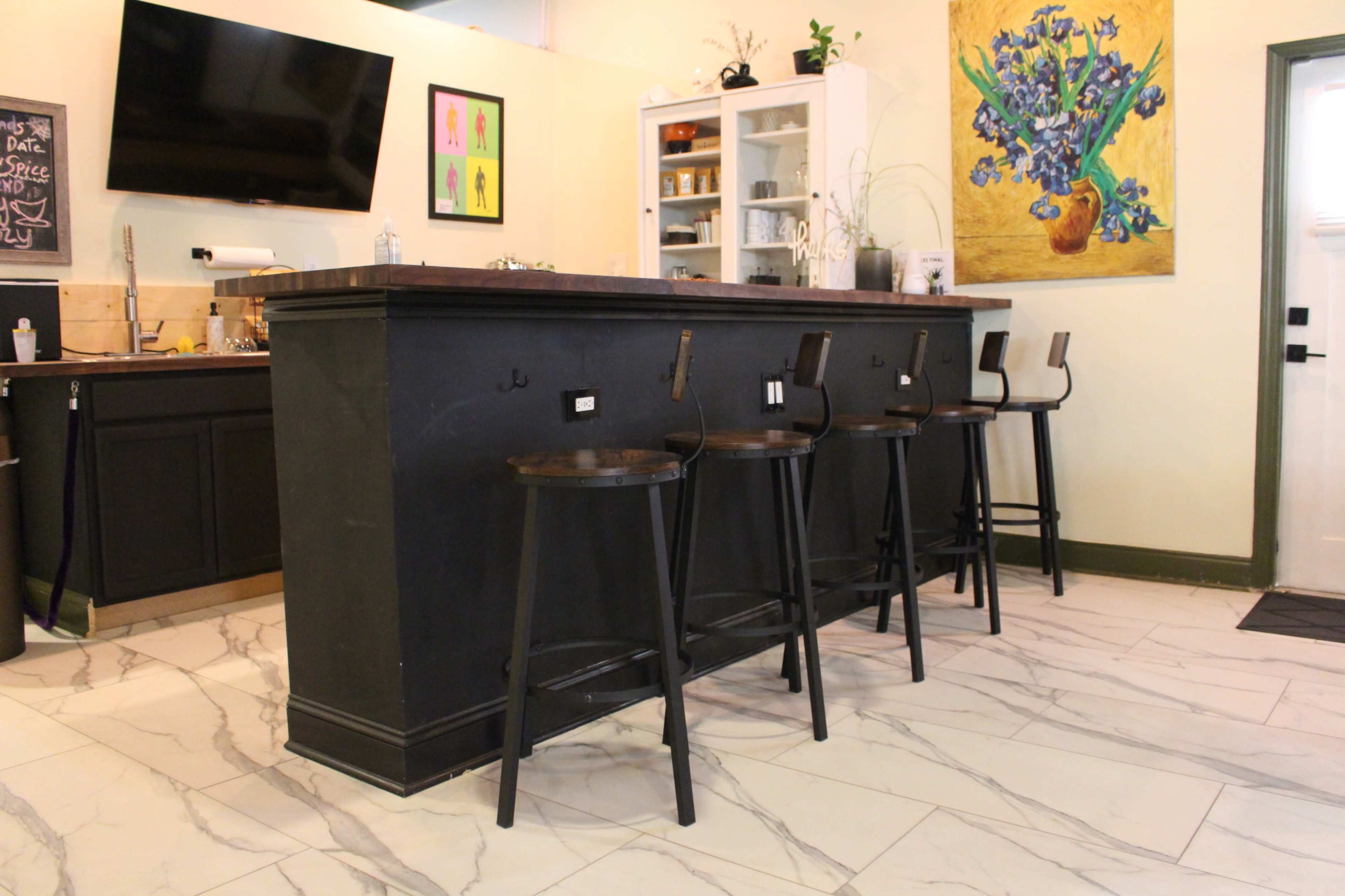 A spacious kitchen island with four black stools is positioned on a marble floor, complemented by a television, a colorful wall painting, and a pantry cabinet in the backdrop.