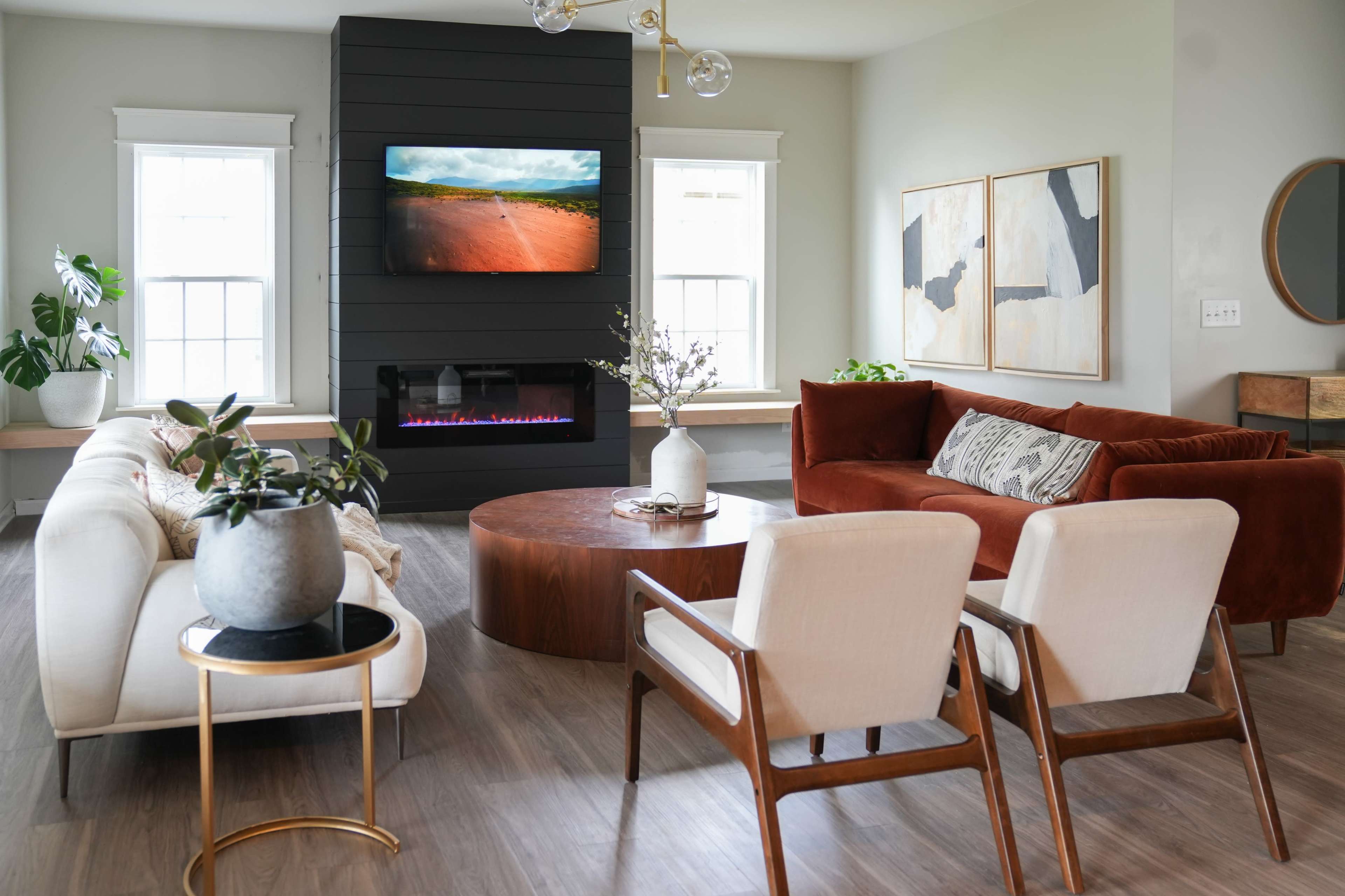 The image shows a modern living room featuring a wall-mounted television above a sleek black fireplace, surrounded by a circular wooden coffee table, two white armchairs, a brown sofa, and decorative plants.