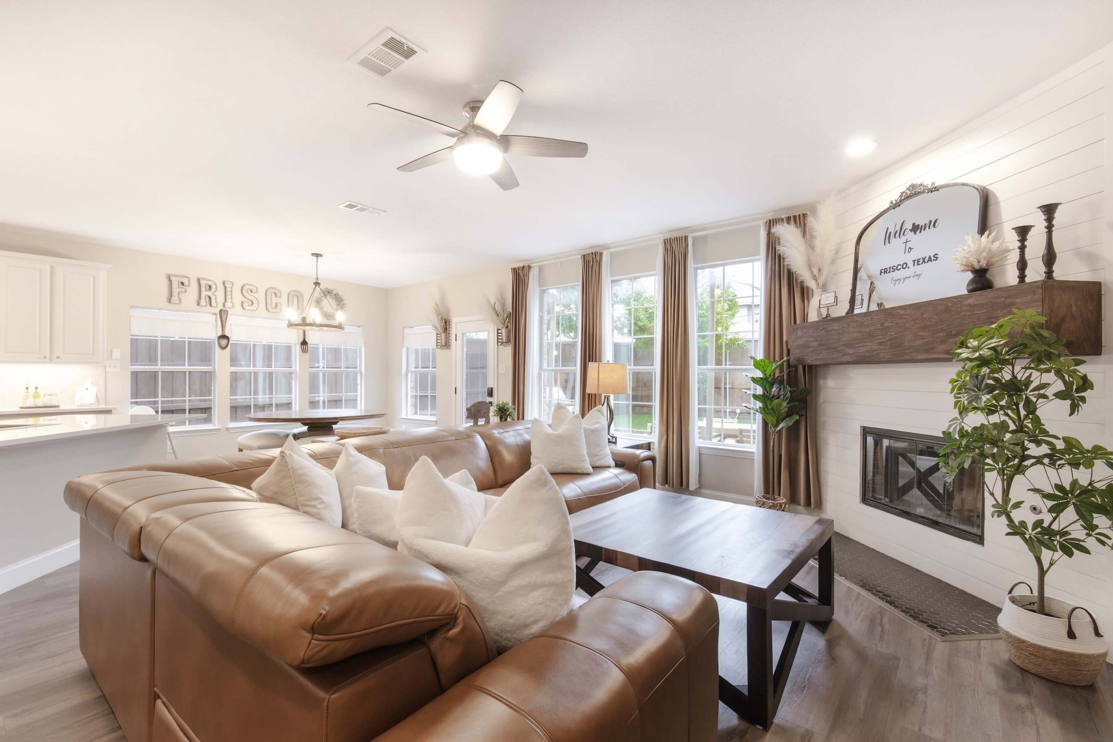 The scene shows a modern living room with a large leather sofa, a wooden coffee table, a fireplace, and windows allowing natural light to fill the space.