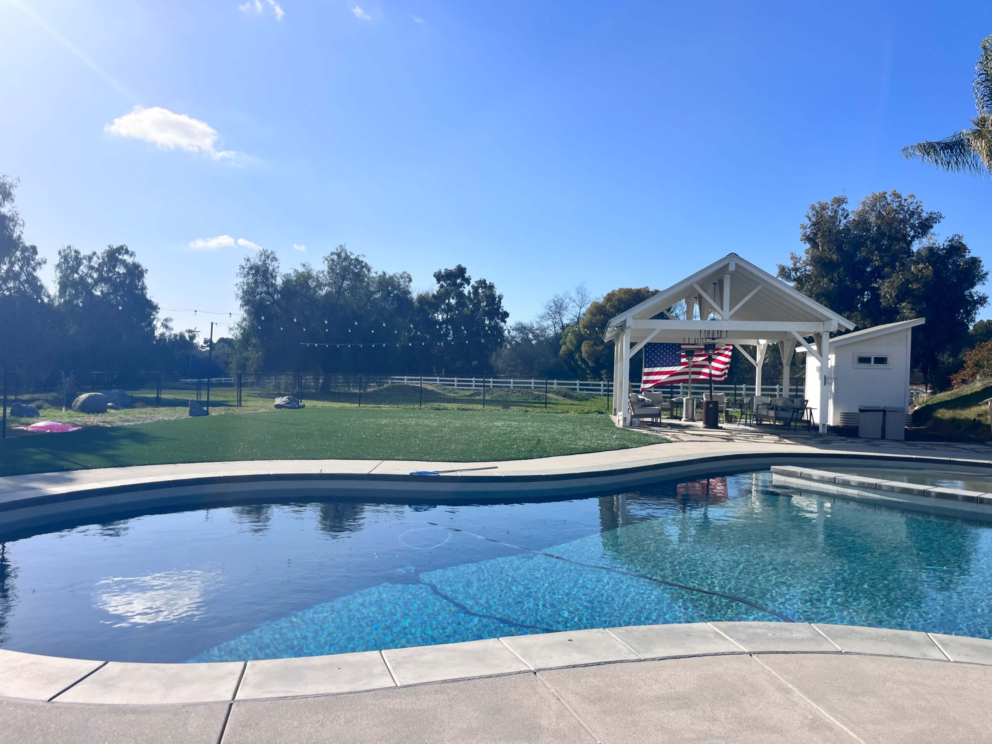 The image shows a swimming pool in the foreground with a covered patio and an American flag in the background, surrounded by greenery and a clear blue sky.
