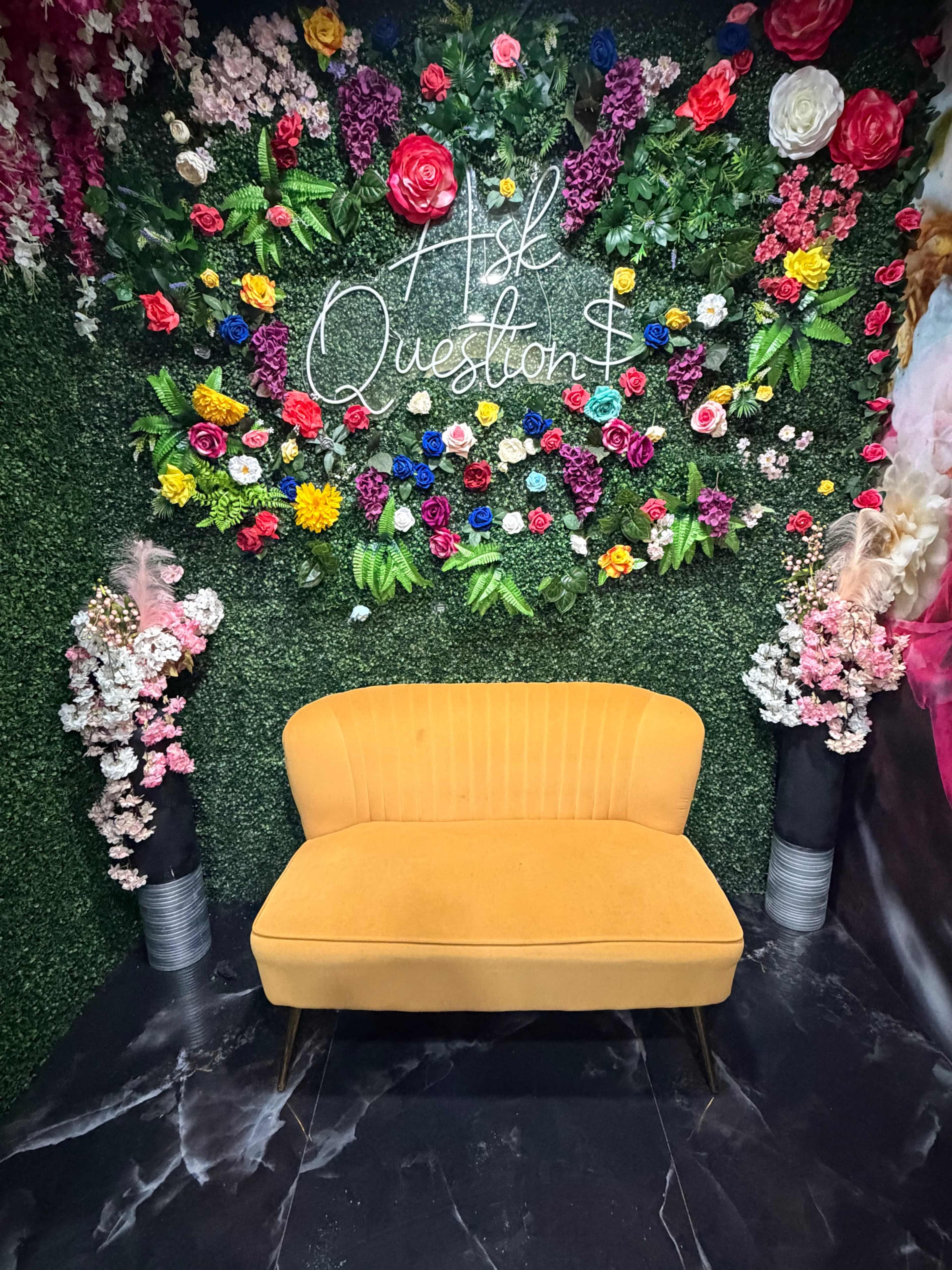 A bright yellow sofa is positioned in front of a floral backdrop decorated with various colorful flowers and greenery.