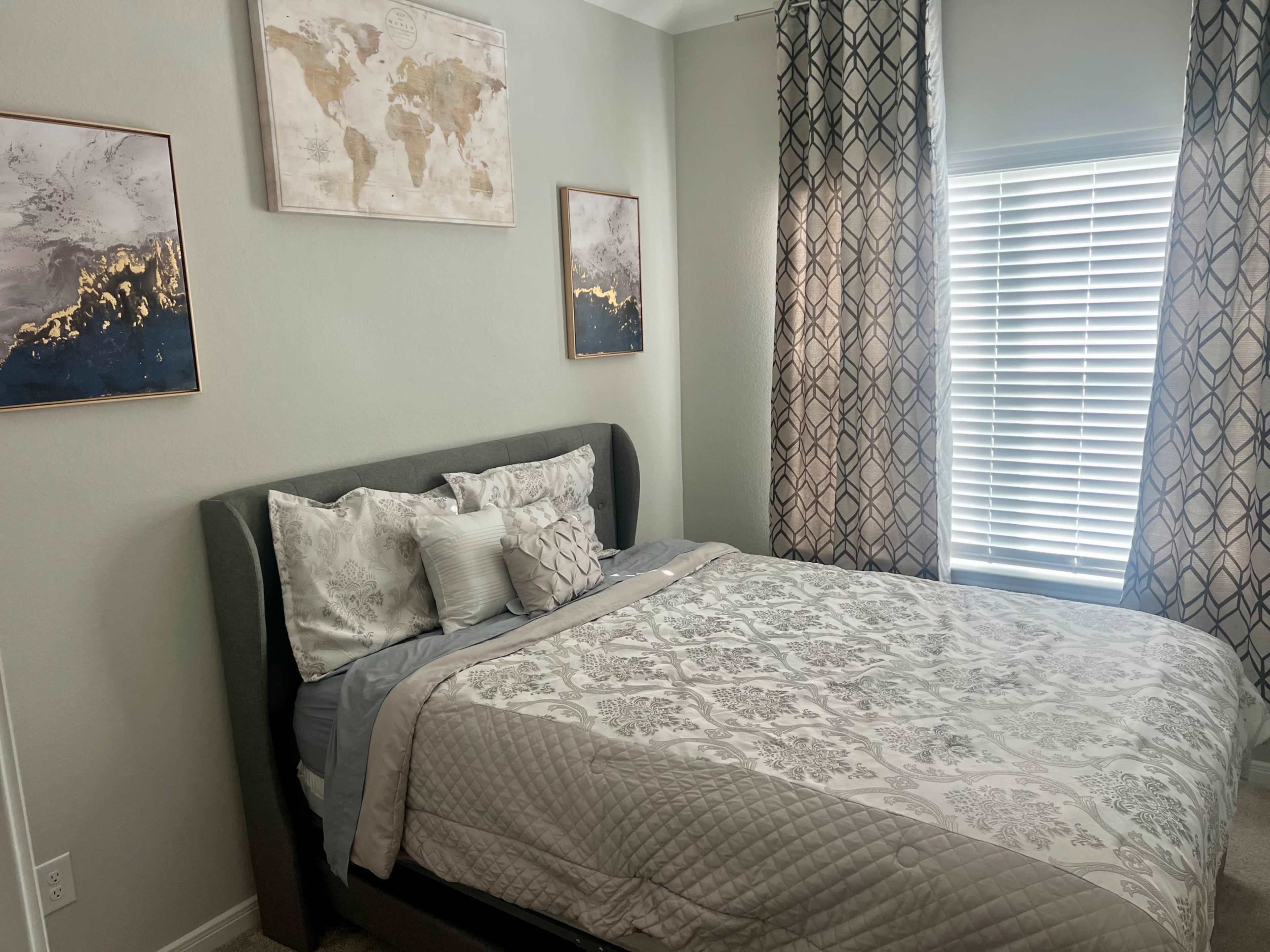 A neatly arranged bedroom featuring a gray upholstered bed with patterned bedding, two wall art pieces, and light-colored curtains.