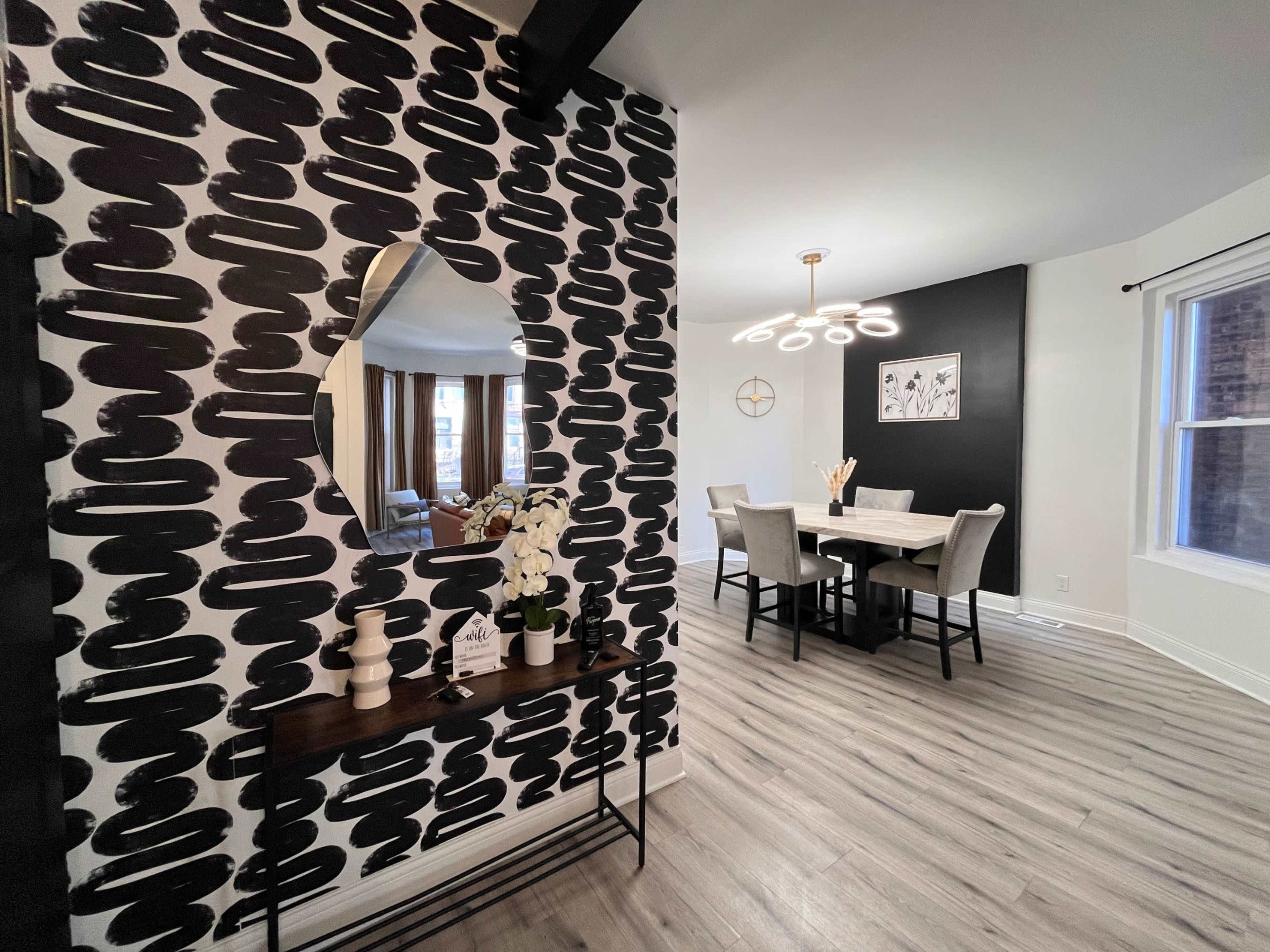 A modern interior with a black and white patterned accent wall, a mirrored decoration, and a dining area featuring a round pendant light and a circular table with gray chairs.