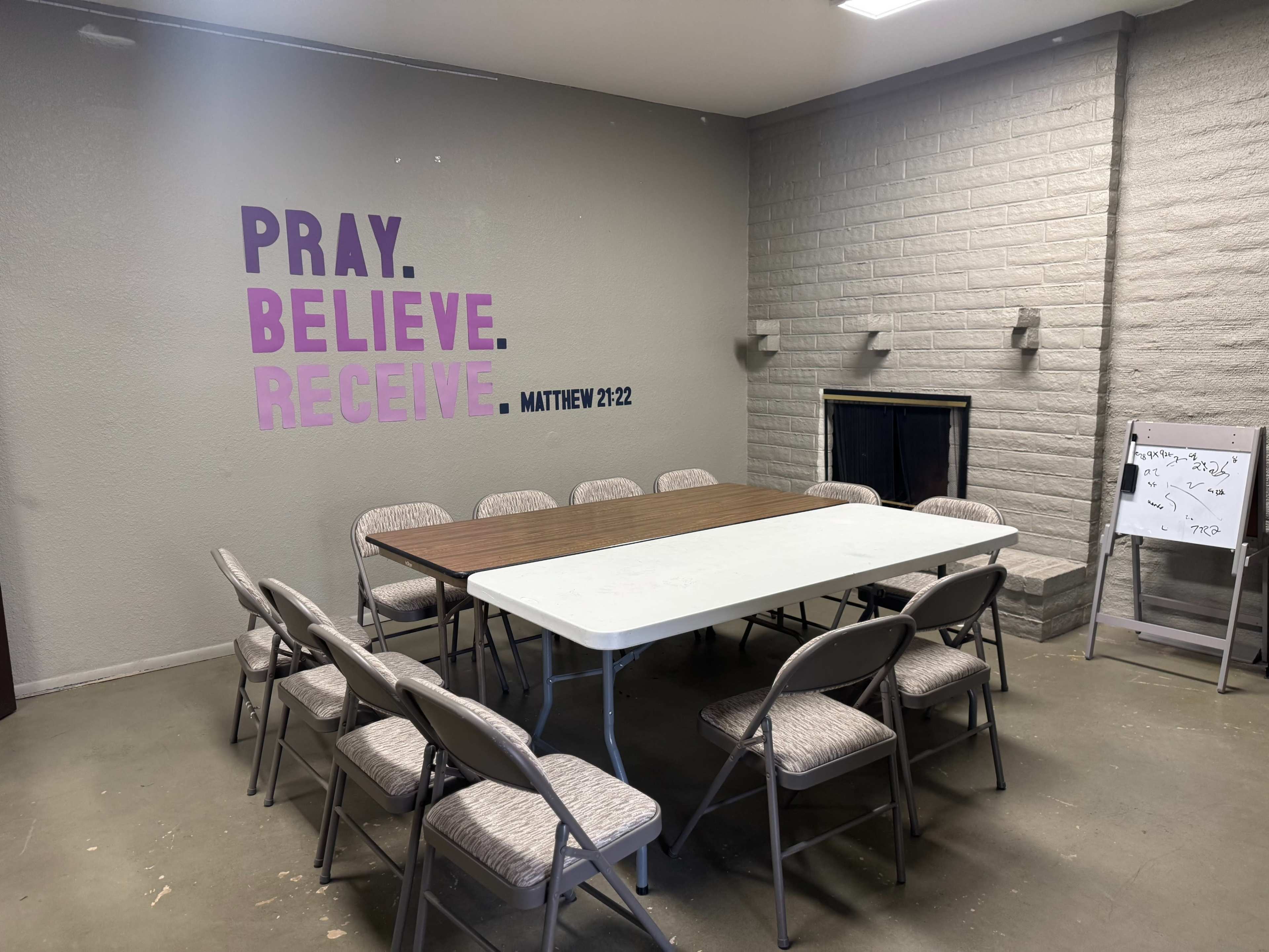 The image shows a meeting room with a rectangular table surrounded by folding chairs and a wall featuring the words "PRAY. BELIEVE. RECEIVE. MATTHEW 21:22."