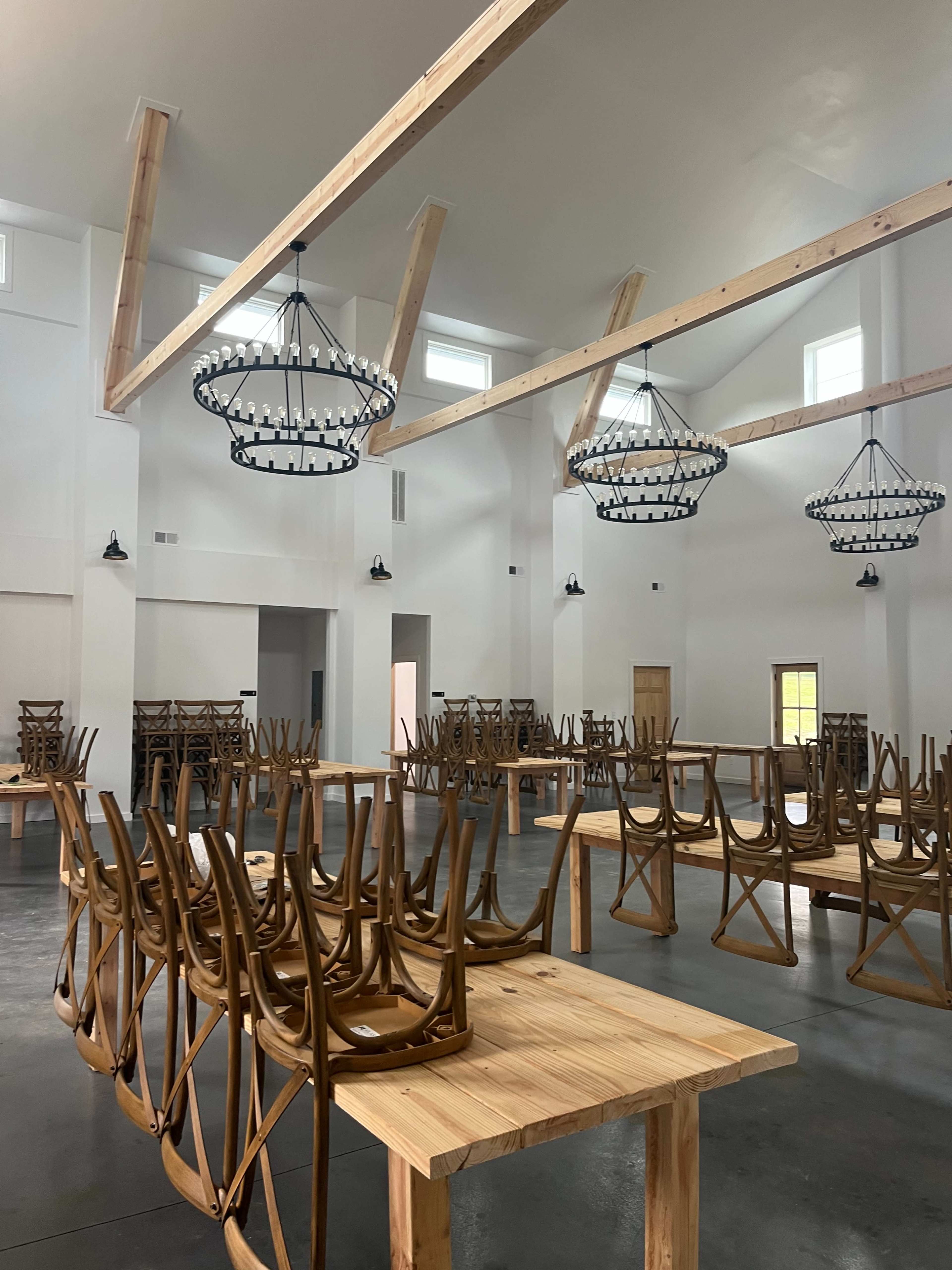 The interior of a spacious, empty room features wooden tables with chairs stacked upside down on top, and large chandeliers hanging from the ceiling.