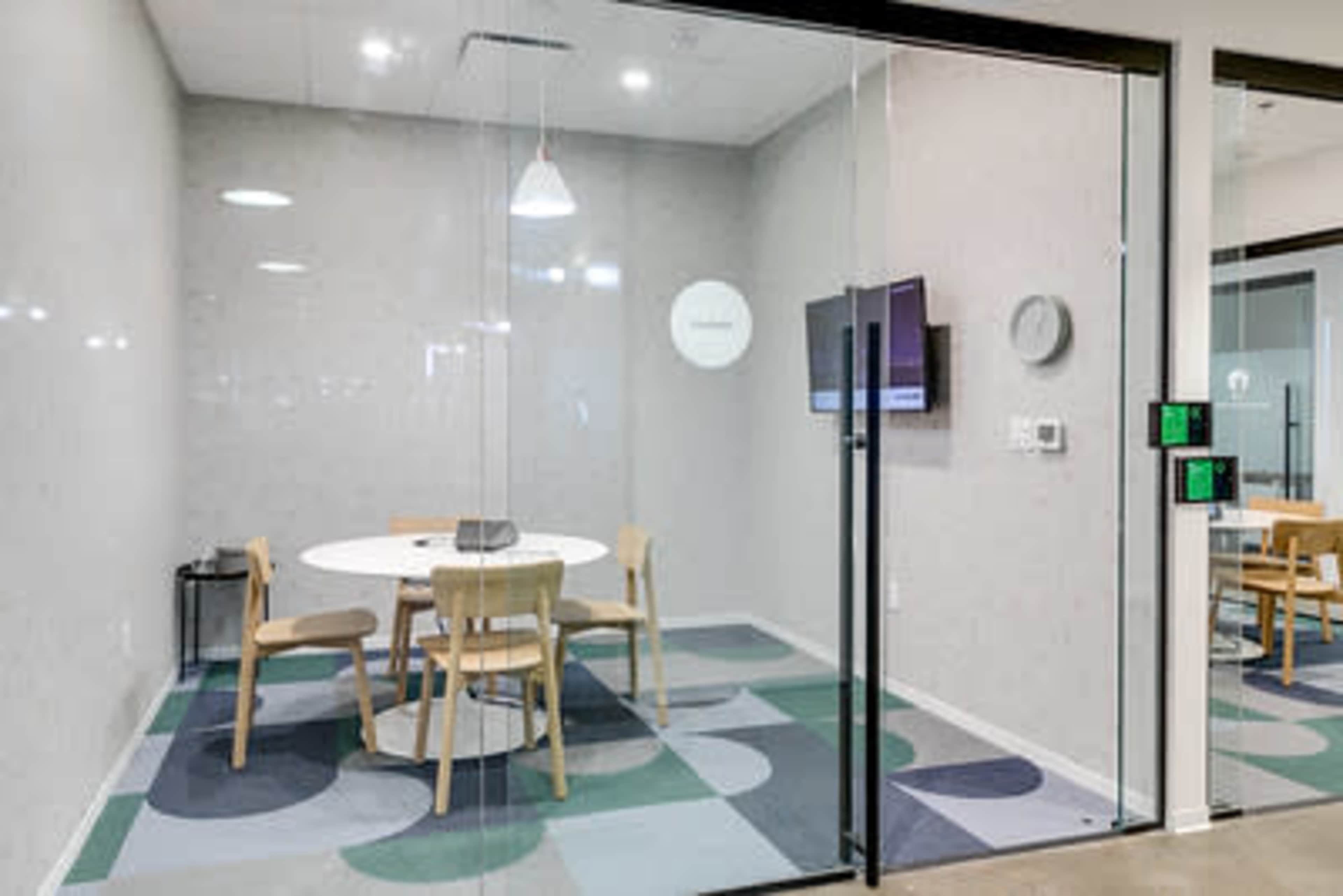 The image shows a small meeting room with a circular table and four chairs, featuring a wall-mounted TV and a clock.