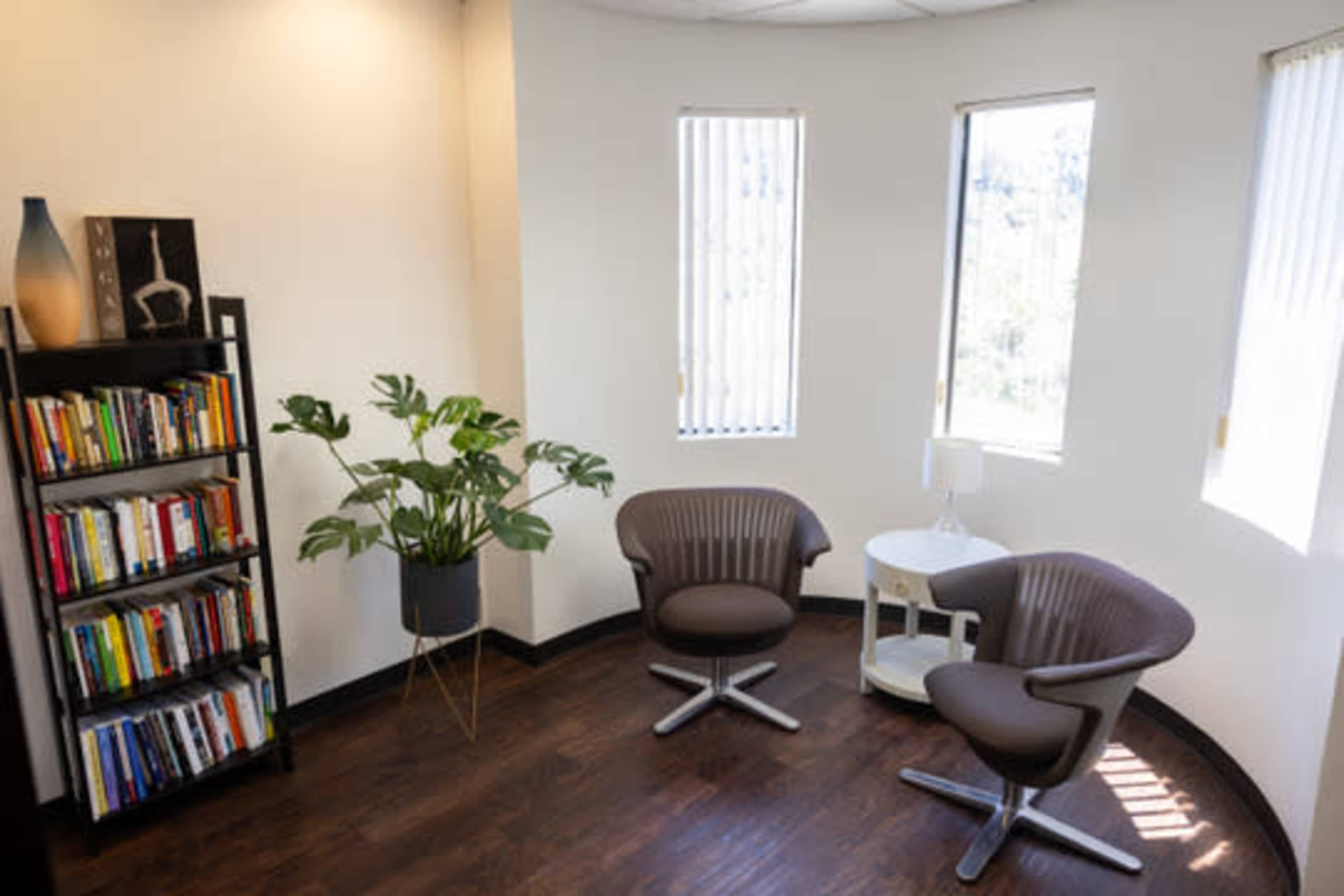 The image shows a cozy reading corner featuring two modern chairs, a small table, a bookshelf filled with books, and a potted plant next to large windows.