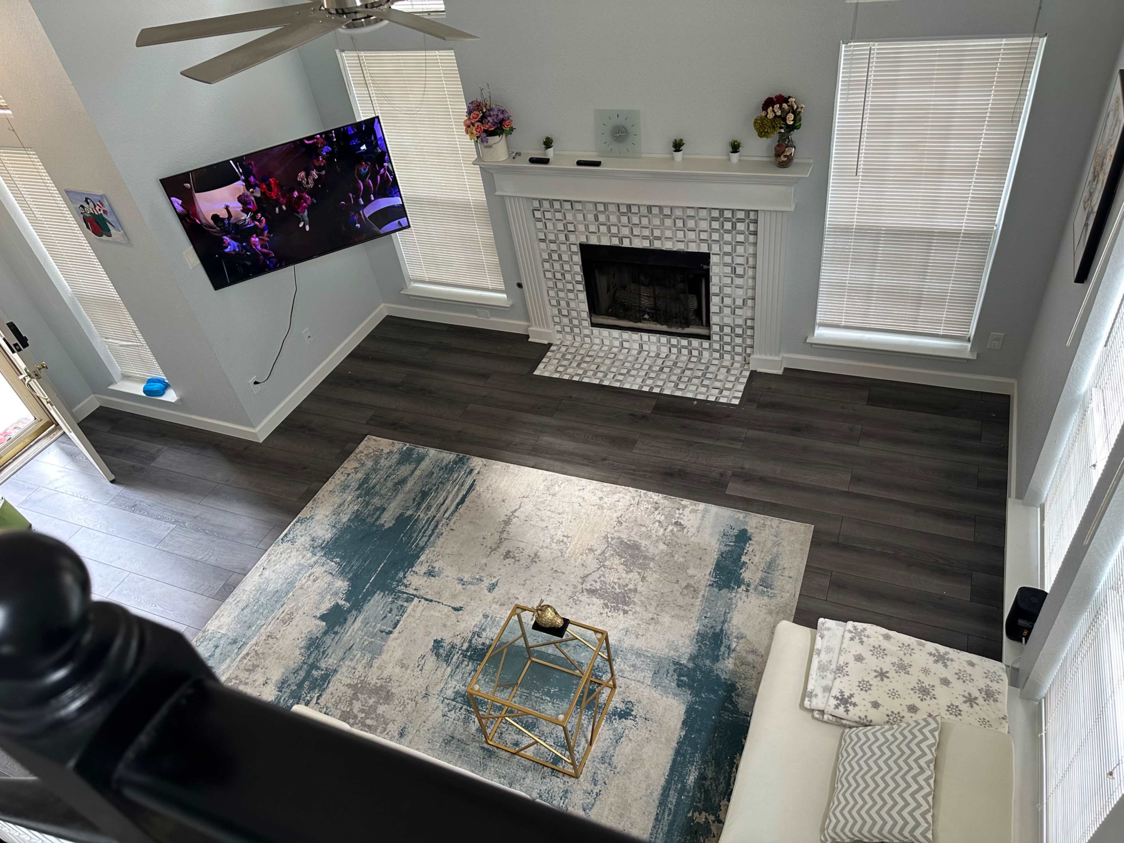 A living room with a television mounted on the wall, a fireplace with a tiled surround, a large area rug on the floor, and a light-colored sofa.