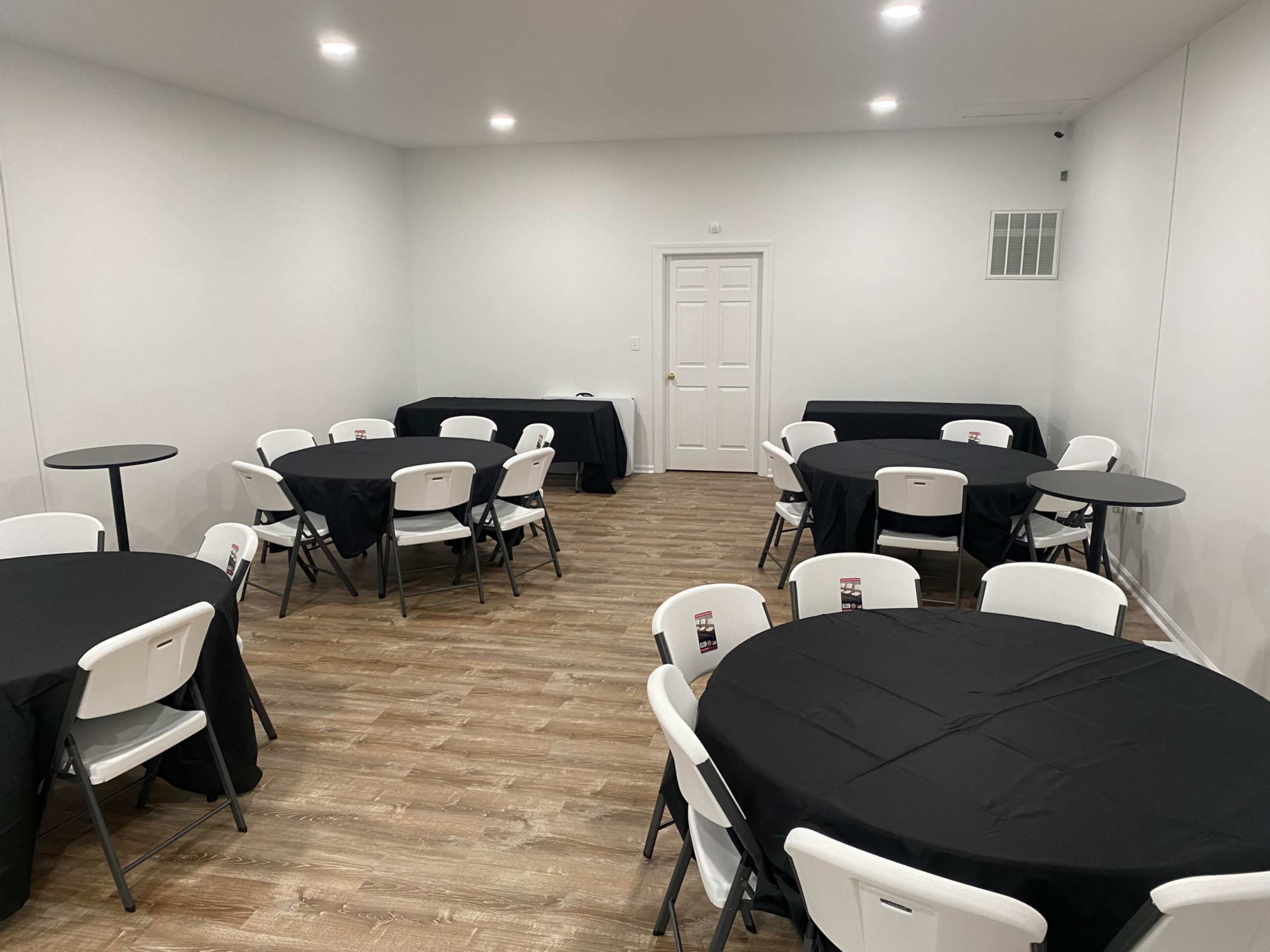 A clean, well-lit event space features several round tables covered with black tablecloths and white chairs arranged around them.