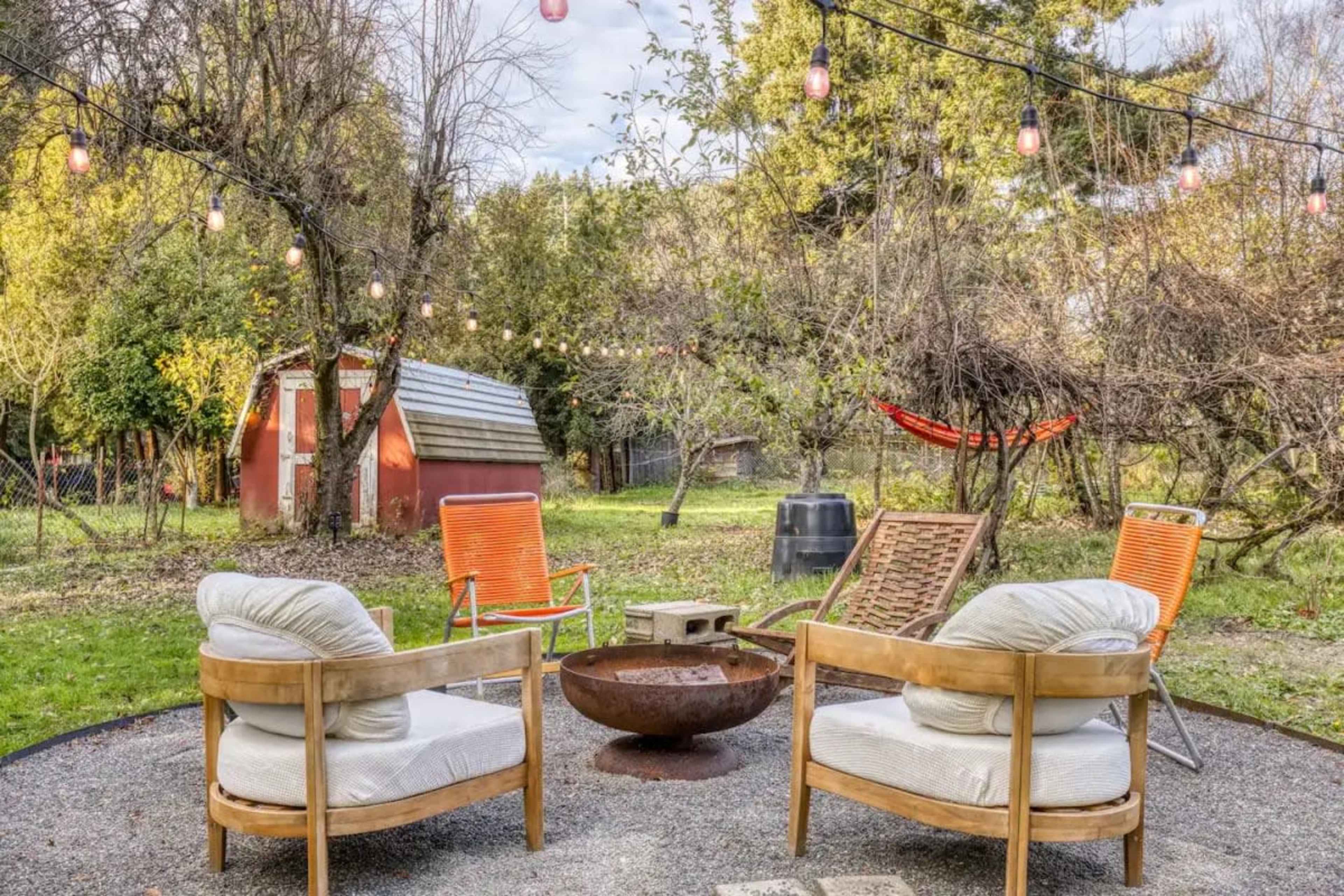The image shows a backyard area with four outdoor chairs arranged around a circular fire pit, string lights overhead, and a red shed in the background.