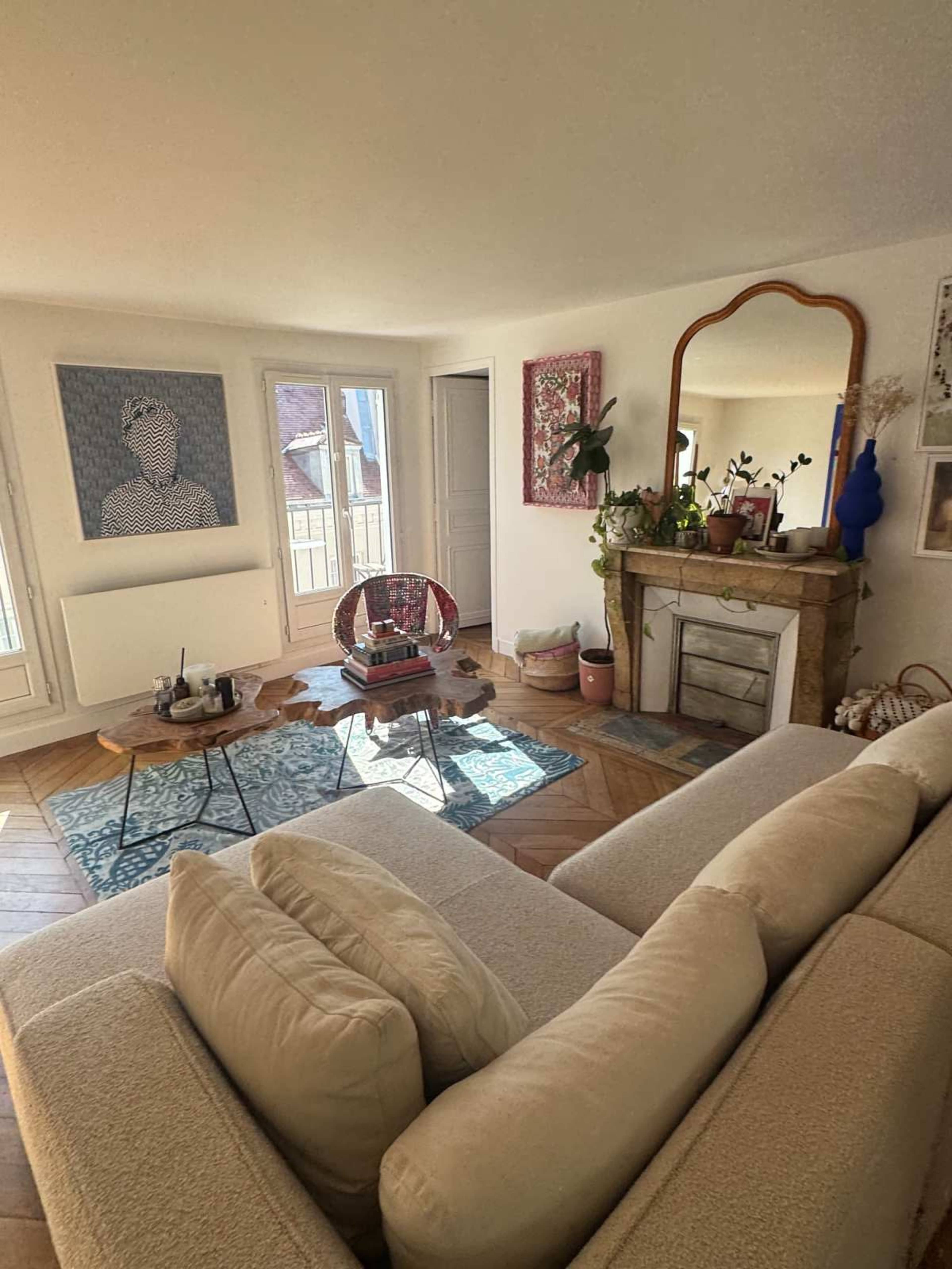 The image shows a bright living room featuring a light-colored sofa, a wooden coffee table, and a decorative fireplace with framed artworks above it.