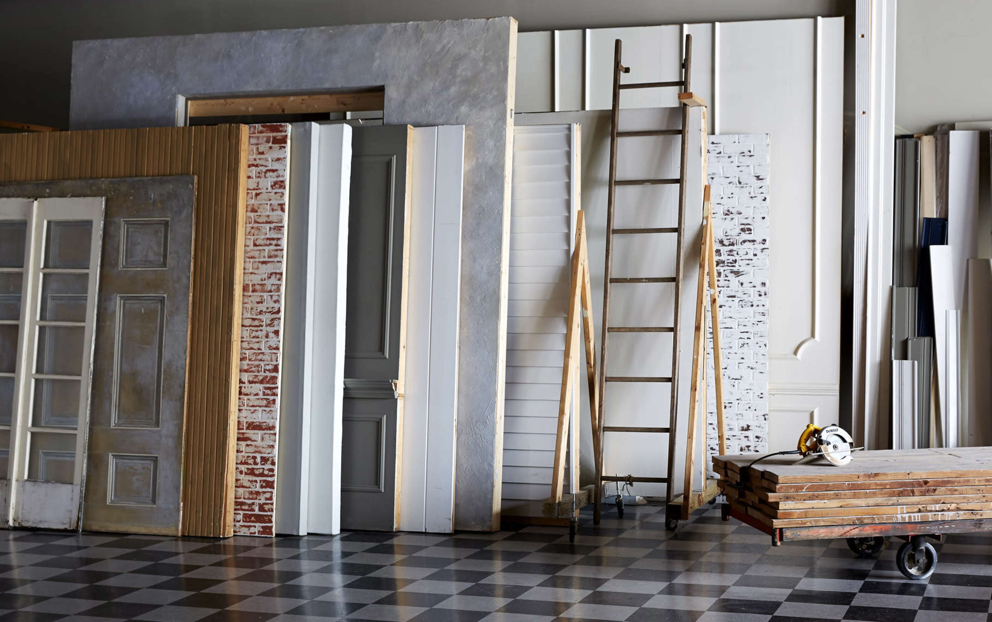 A collection of various doors and wall panels is arranged next to a wooden ladder and a stack of wooden boards in a workshop.