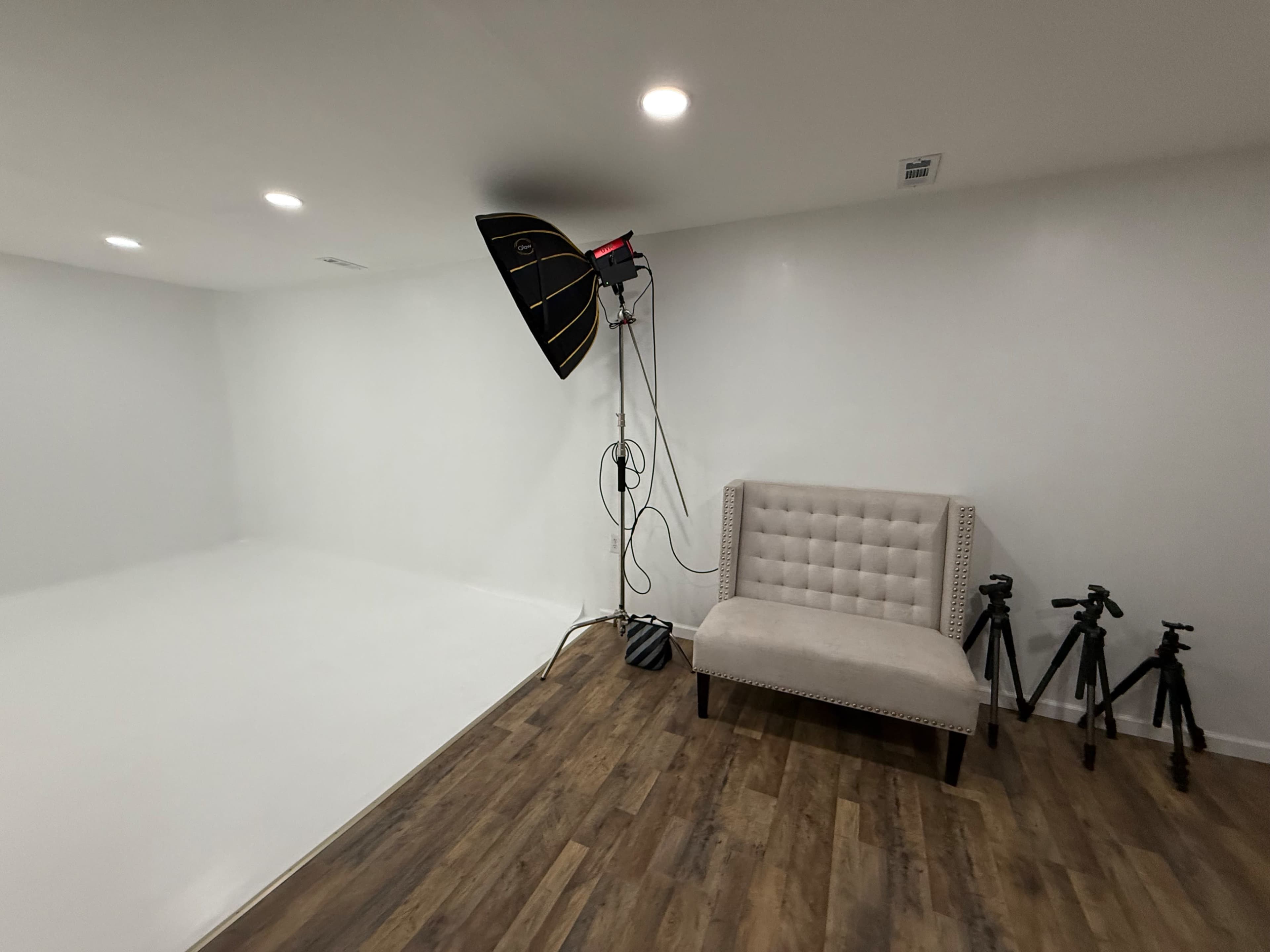 The image shows a studio space with a beige tufted couch, three tripods, and a large softbox lighting setup against a plain white backdrop.
