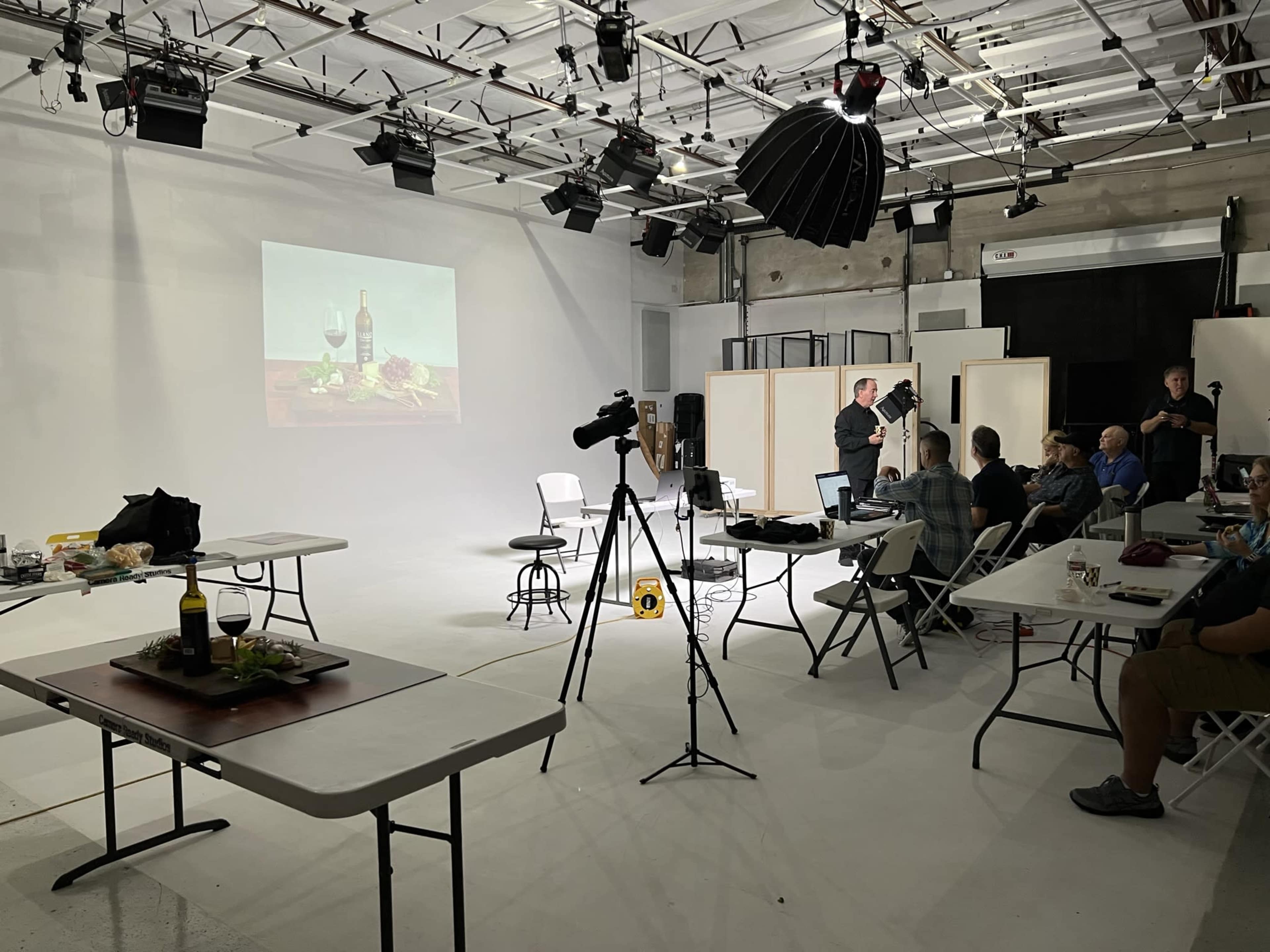 A group of people is gathered in a studio for a photography workshop, with a presentation on food and wine set up on a screen.