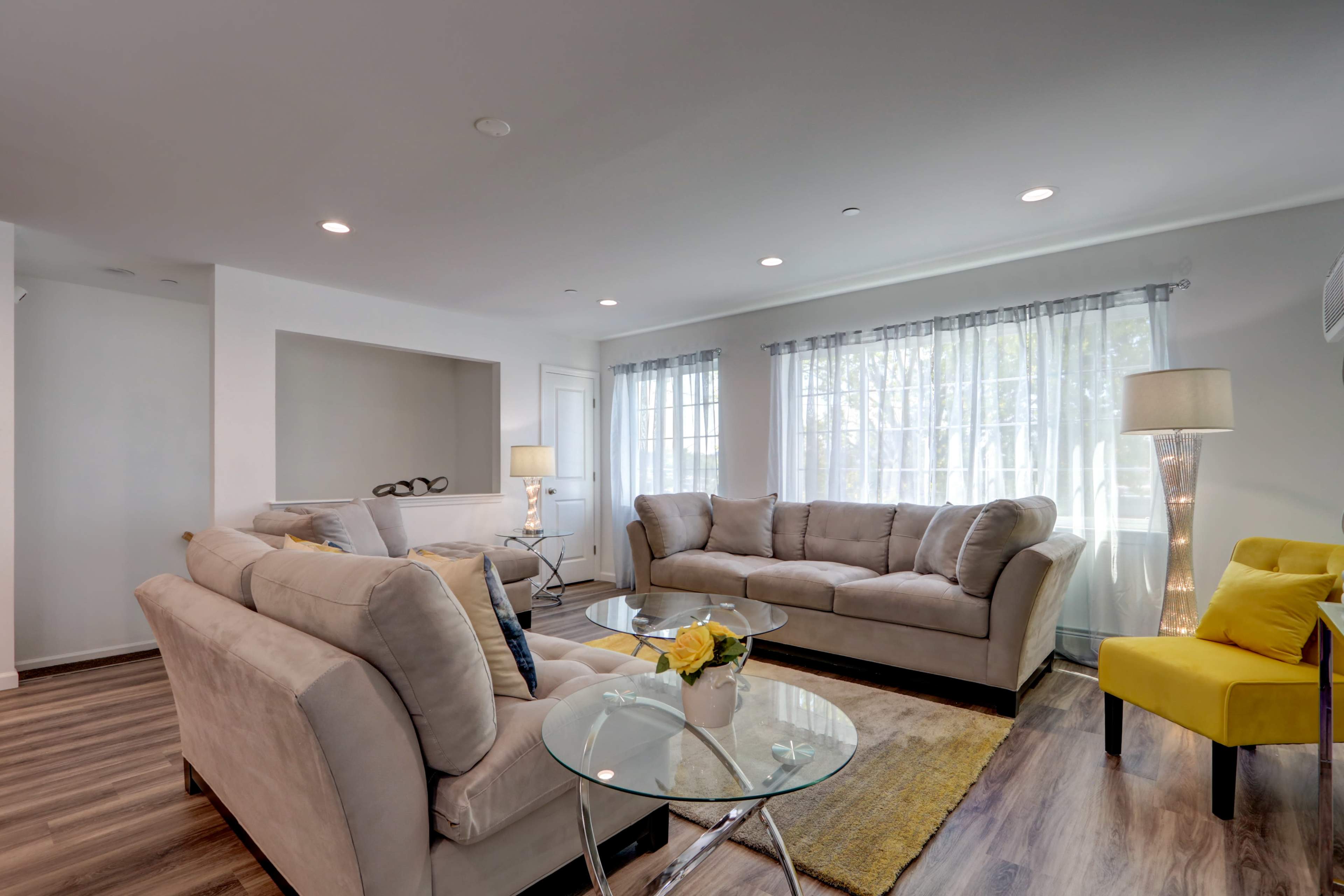 A modern living room with two beige sofas, a yellow armchair, a glass coffee table, and large windows with sheer curtains.