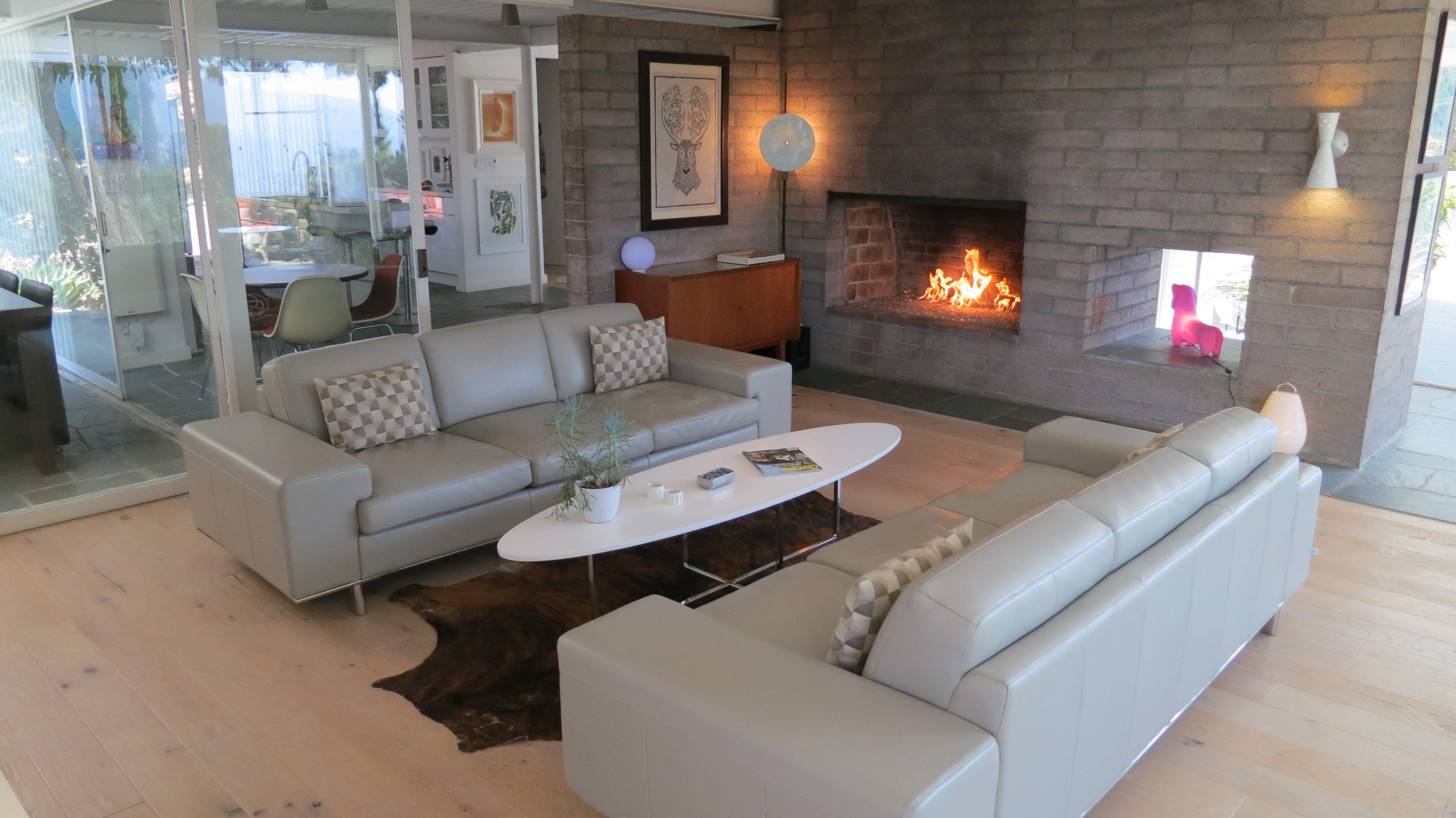 The scene shows a modern living room with two leather sofas facing a stone fireplace, surrounded by large windows and minimalist decor.