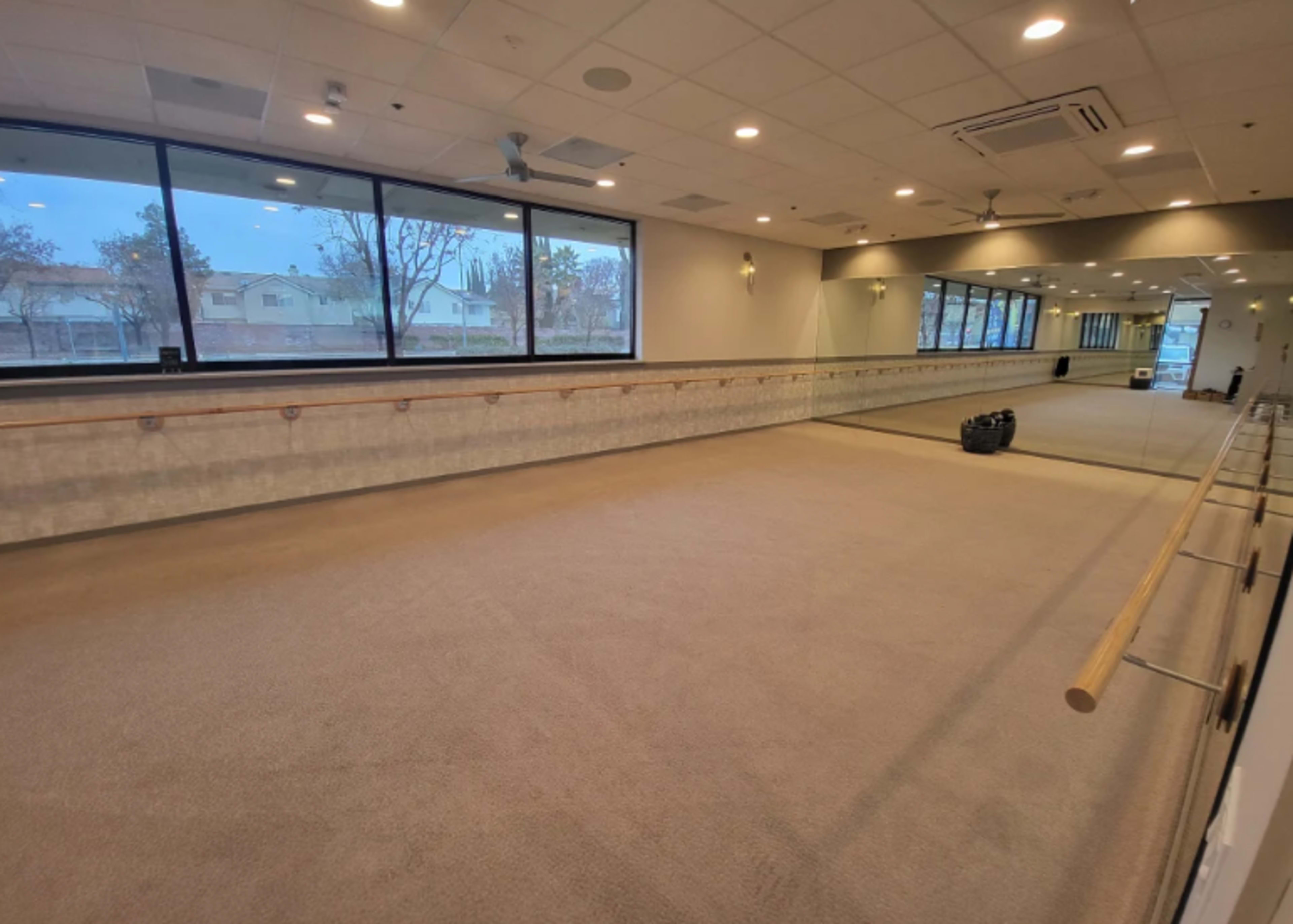 The image shows a spacious workout studio with large windows, a mirrored wall, and a wooden ballet barre along one side.