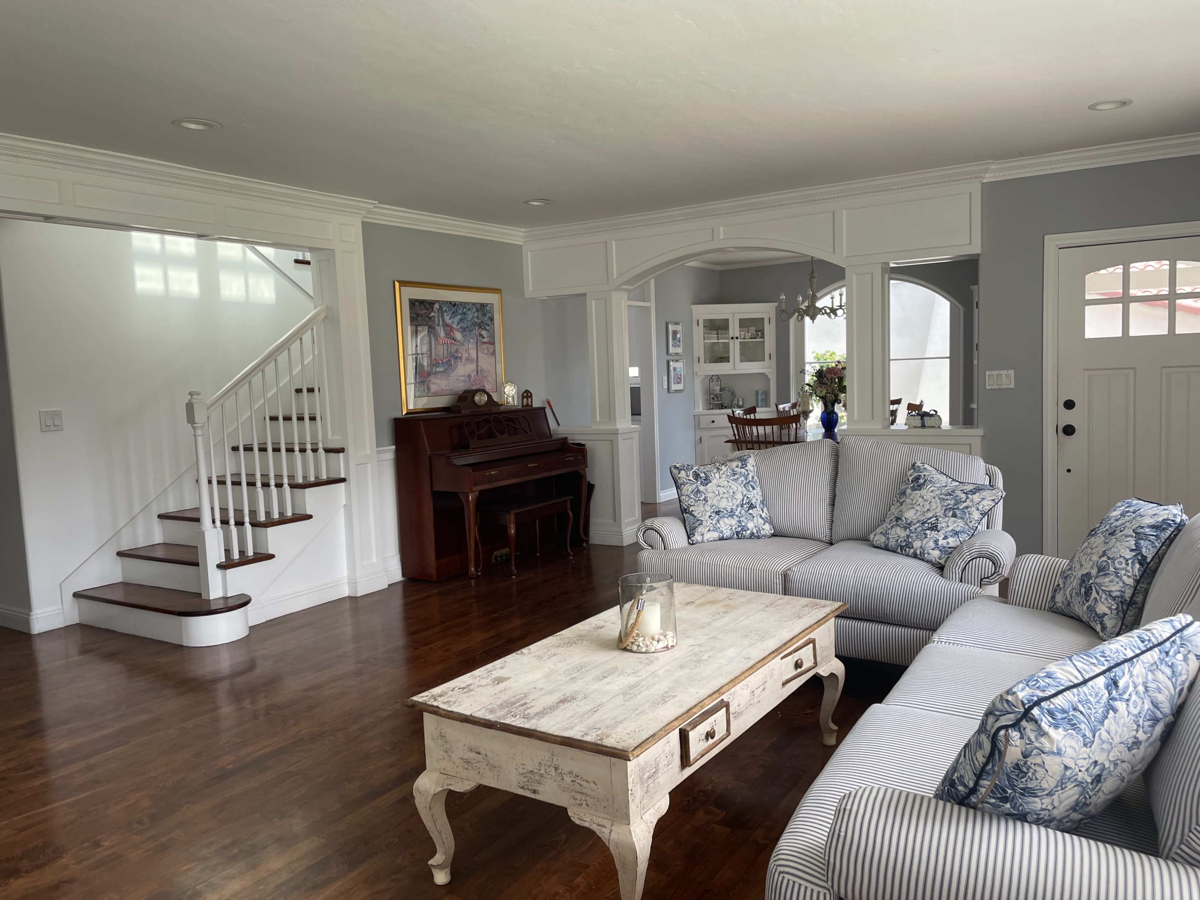 A spacious living room with a gray color scheme, featuring a couch with blue-patterned cushions, a wooden coffee table, a piano in the corner, and a staircase leading to an upper level.