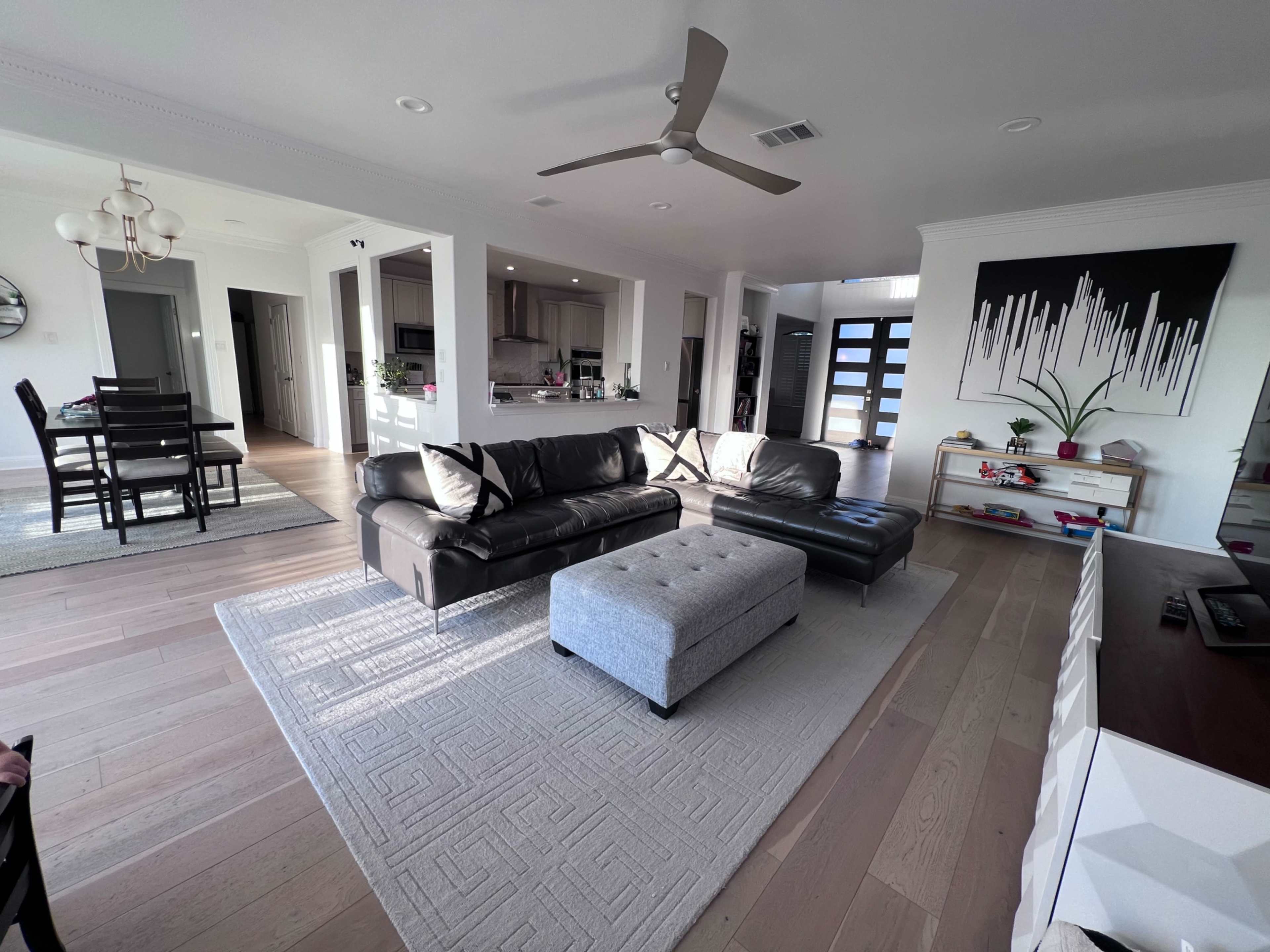 The living room features a black sectional sofa, an ottoman, and a light-colored area rug, with an open layout leading to a kitchen in the background.