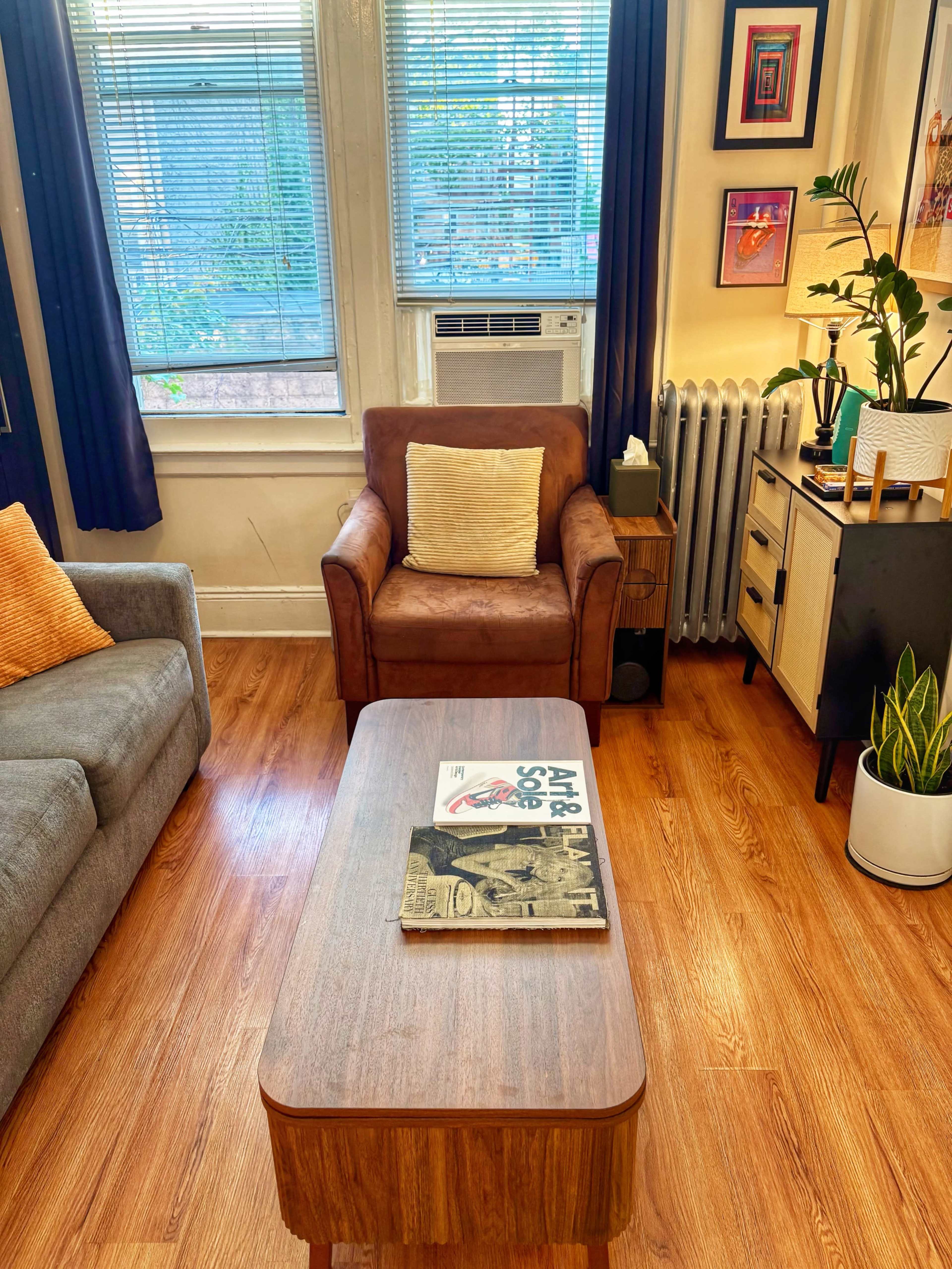 The image shows a living room with a brown armchair, a gray sofa, a wooden coffee table with a magazine, a potted plant, and various wall decorations.