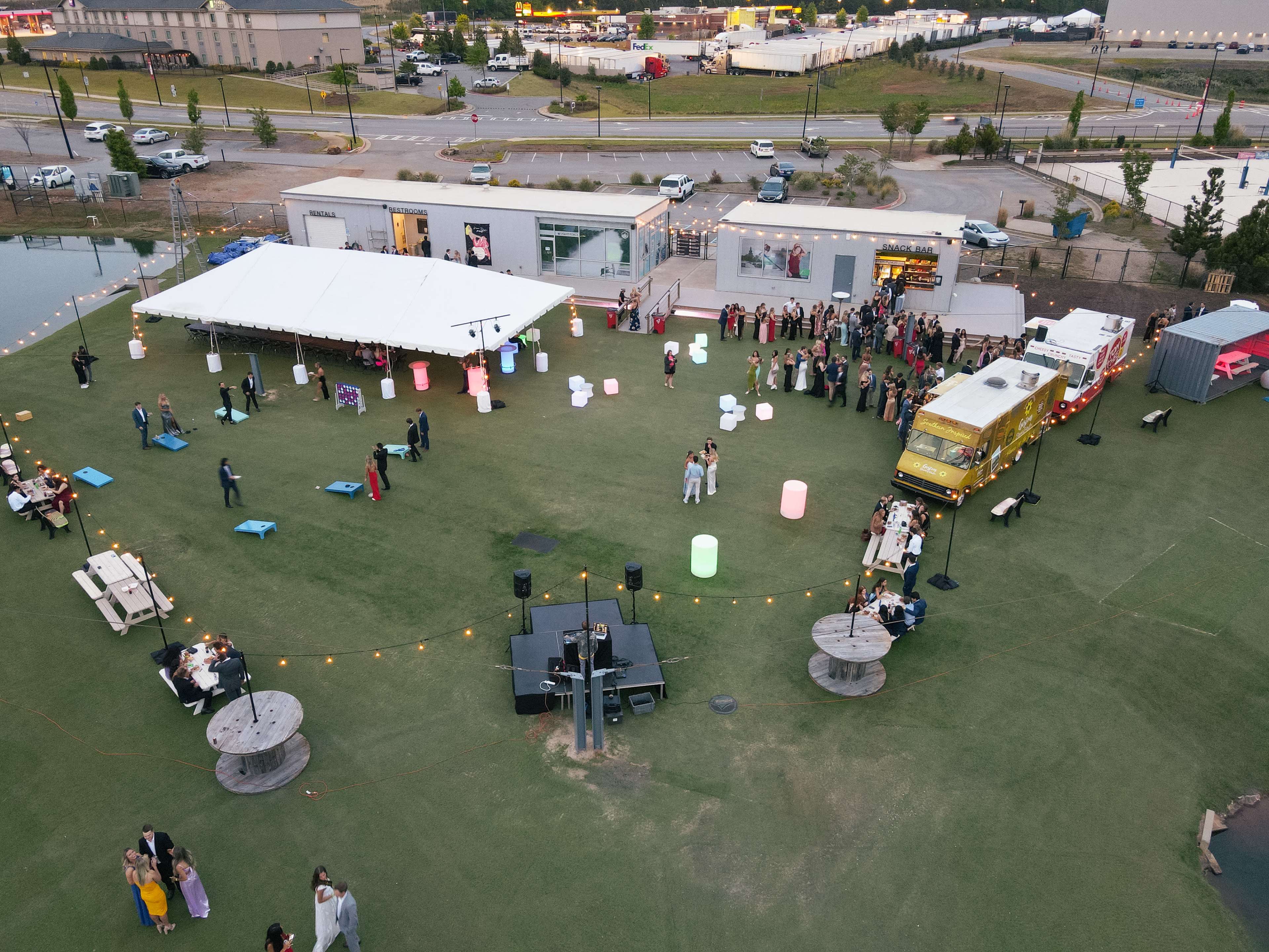 An outdoor event is taking place on a large grassy area with a stage, food truck, and guests mingling near illuminated decorations.
