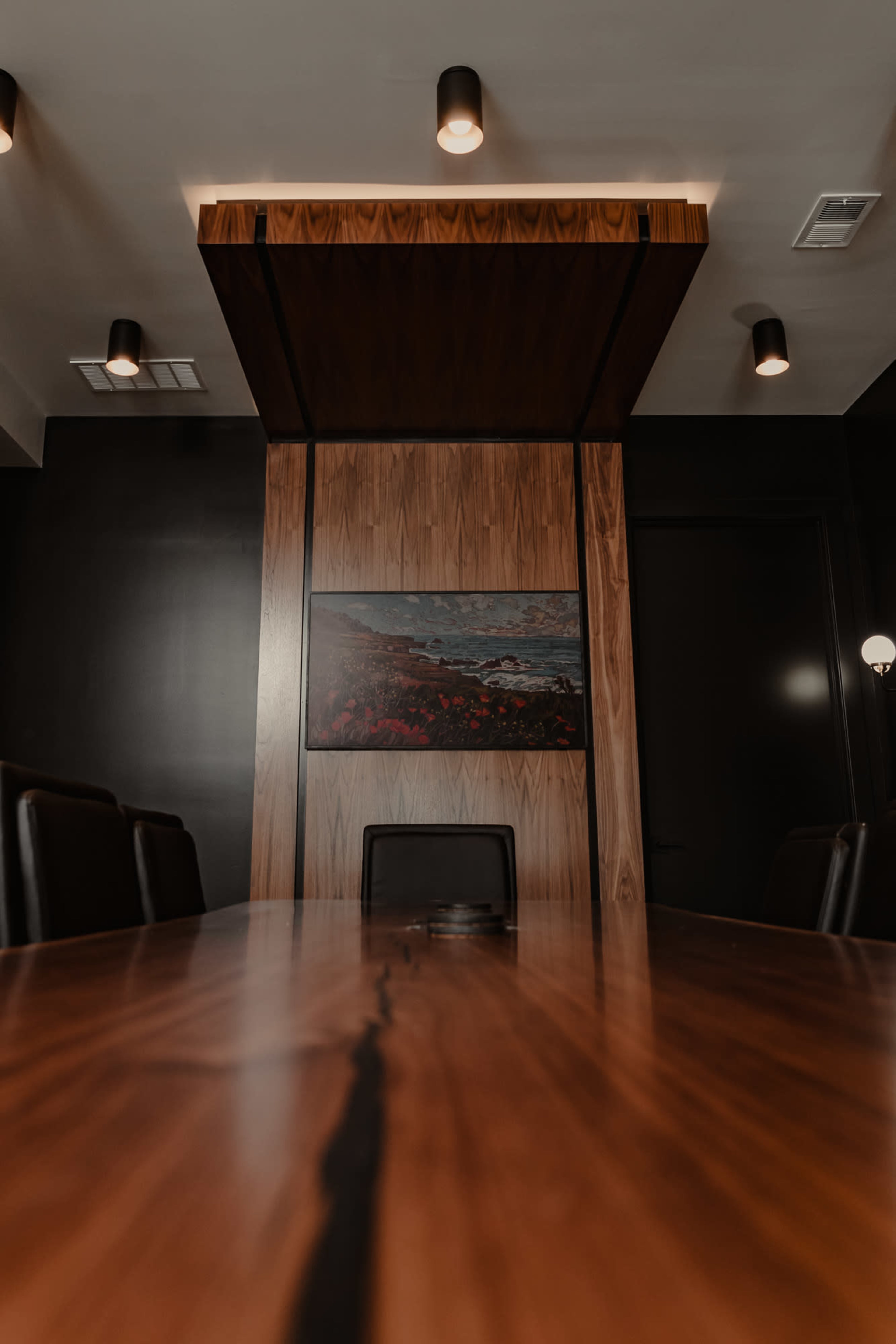 A wooden conference table is centered in a dimly lit room with dark walls and a large painting on the back wall.