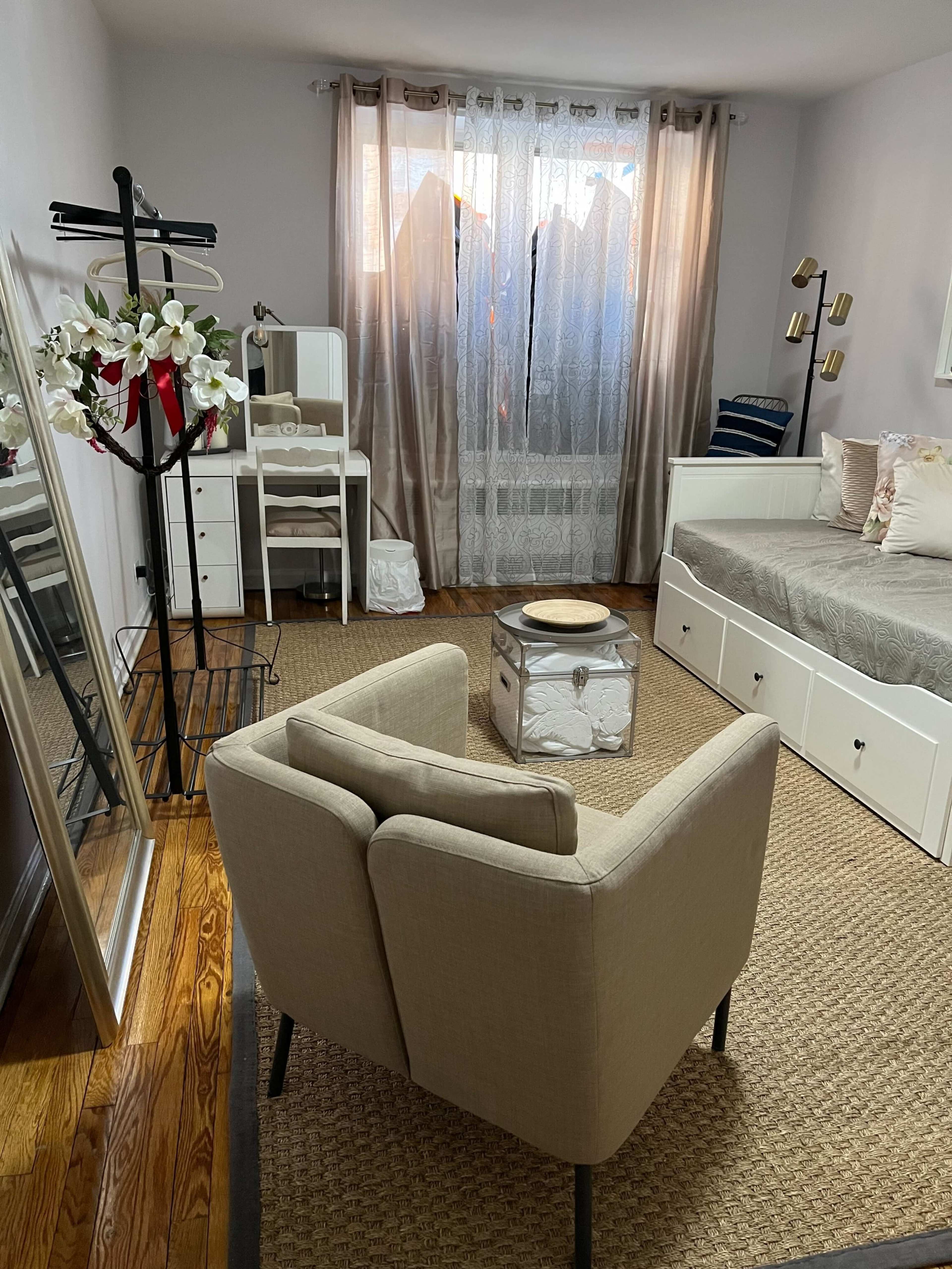 The image shows a cozy room featuring a bed, a desk, a mirror, and a chair, with light curtains and wooden flooring.