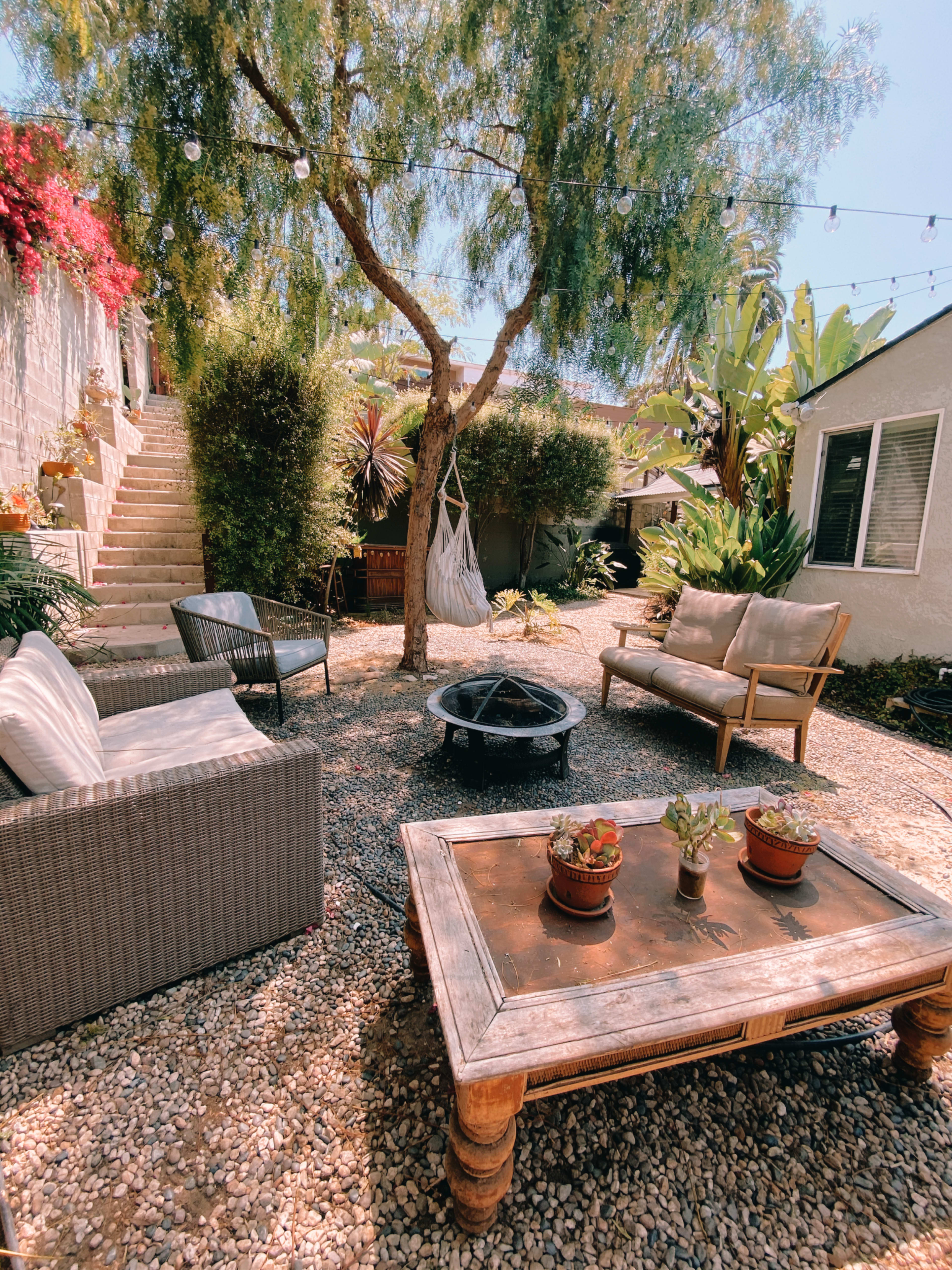 A cozy outdoor space features a fire pit surrounded by seating, a table with potted plants, and a tree amidst gravel and stairs leading to a higher level.
