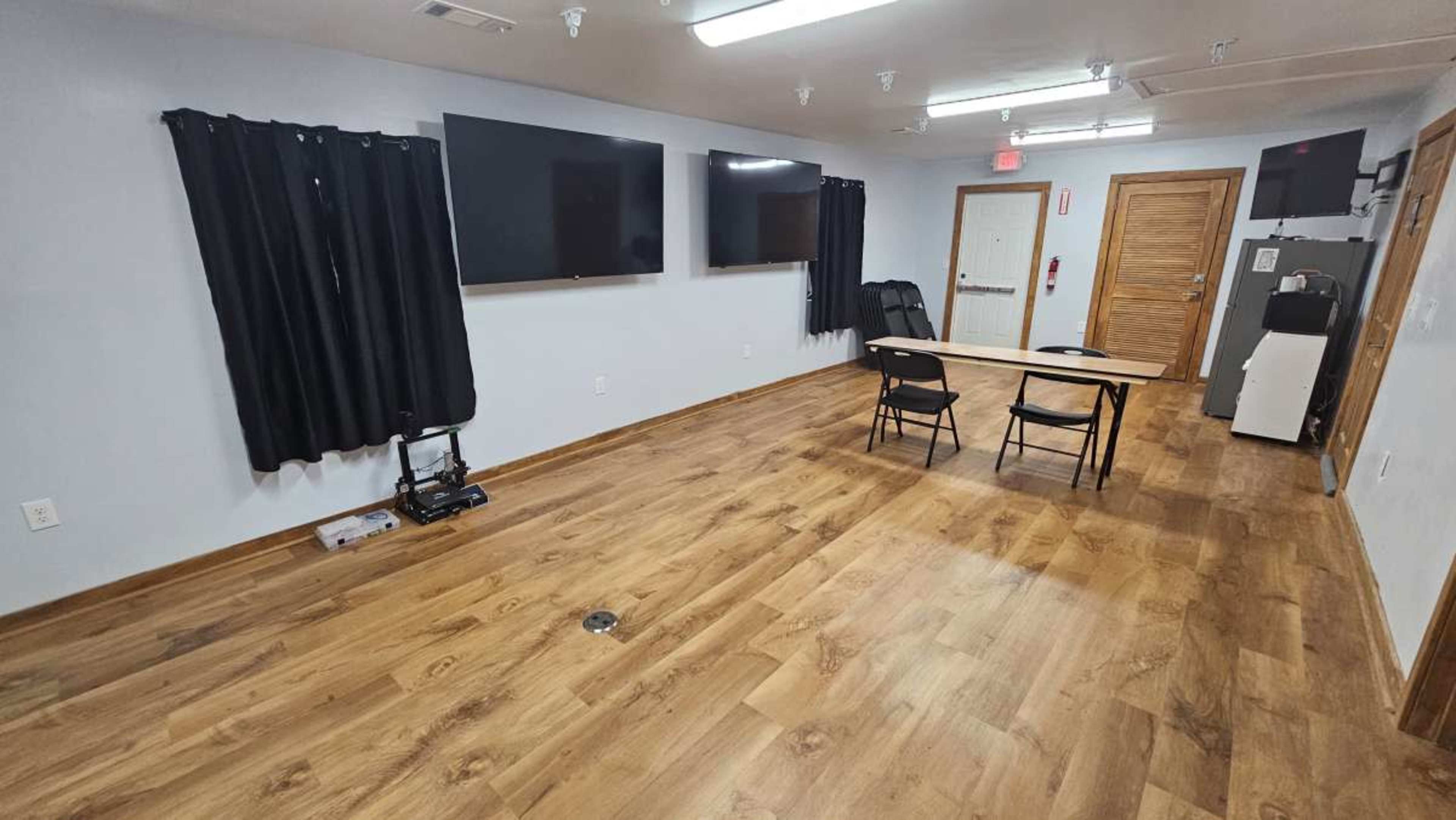 The image shows a room with dark curtains, two large wall-mounted screens, a table with two chairs, and a kitchenette area against one wall.