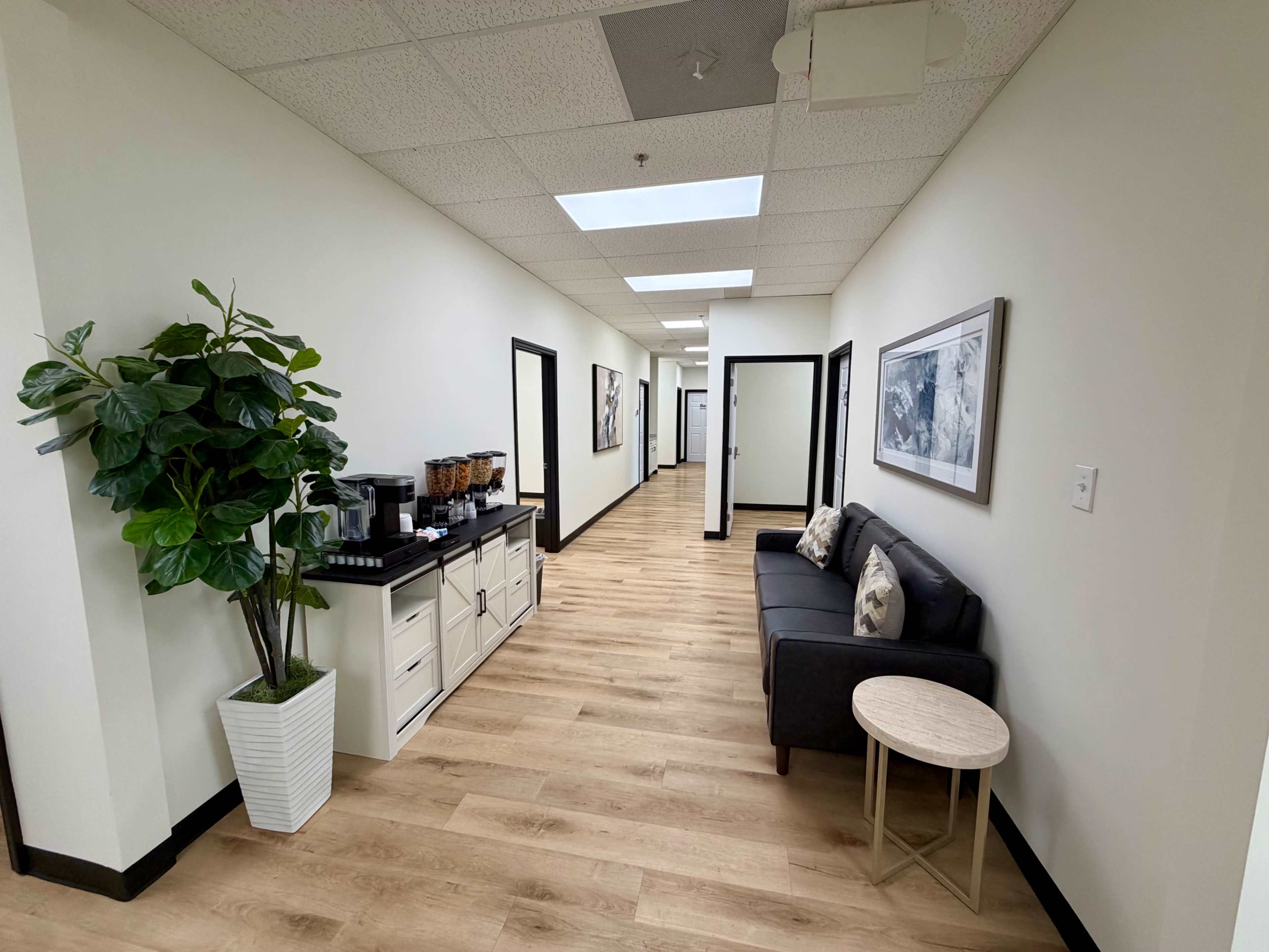 The image shows a modern hallway with a coffee station, a seating area featuring a gray sofa and a small table, and several doorways on either side.