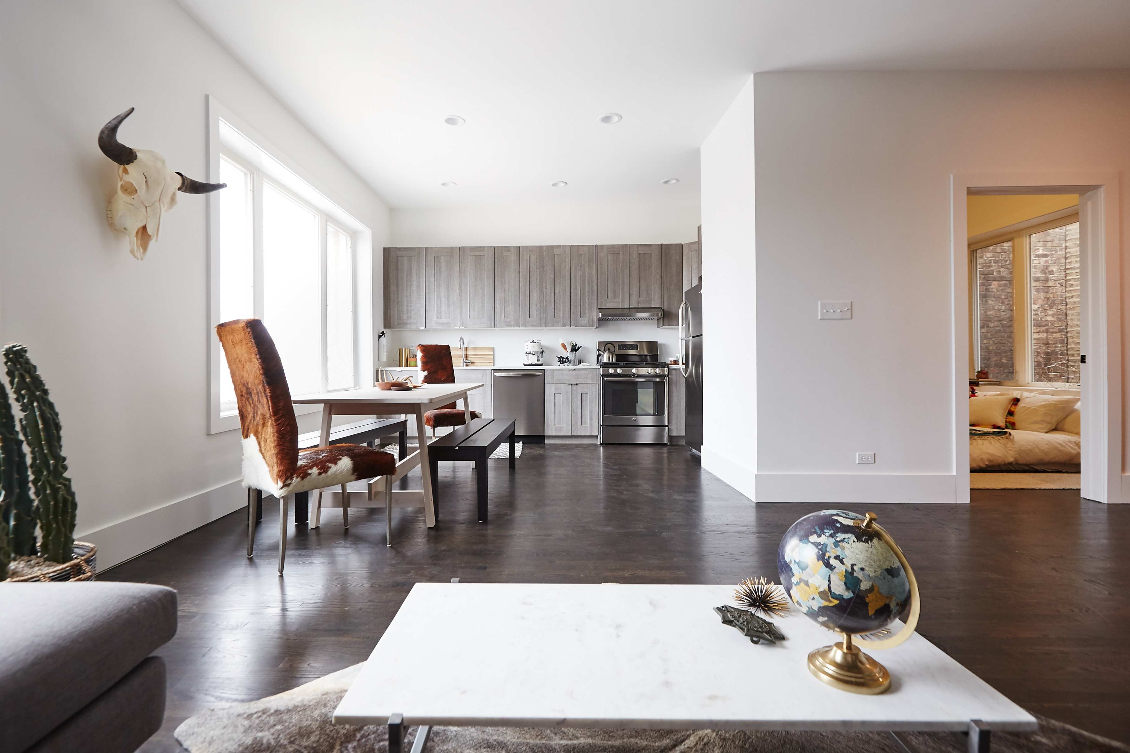 A modern kitchen and dining area features a wooden table, leather chairs, and stainless steel appliances, with a living space showcasing a globe on a coffee table and a decorative wall piece.