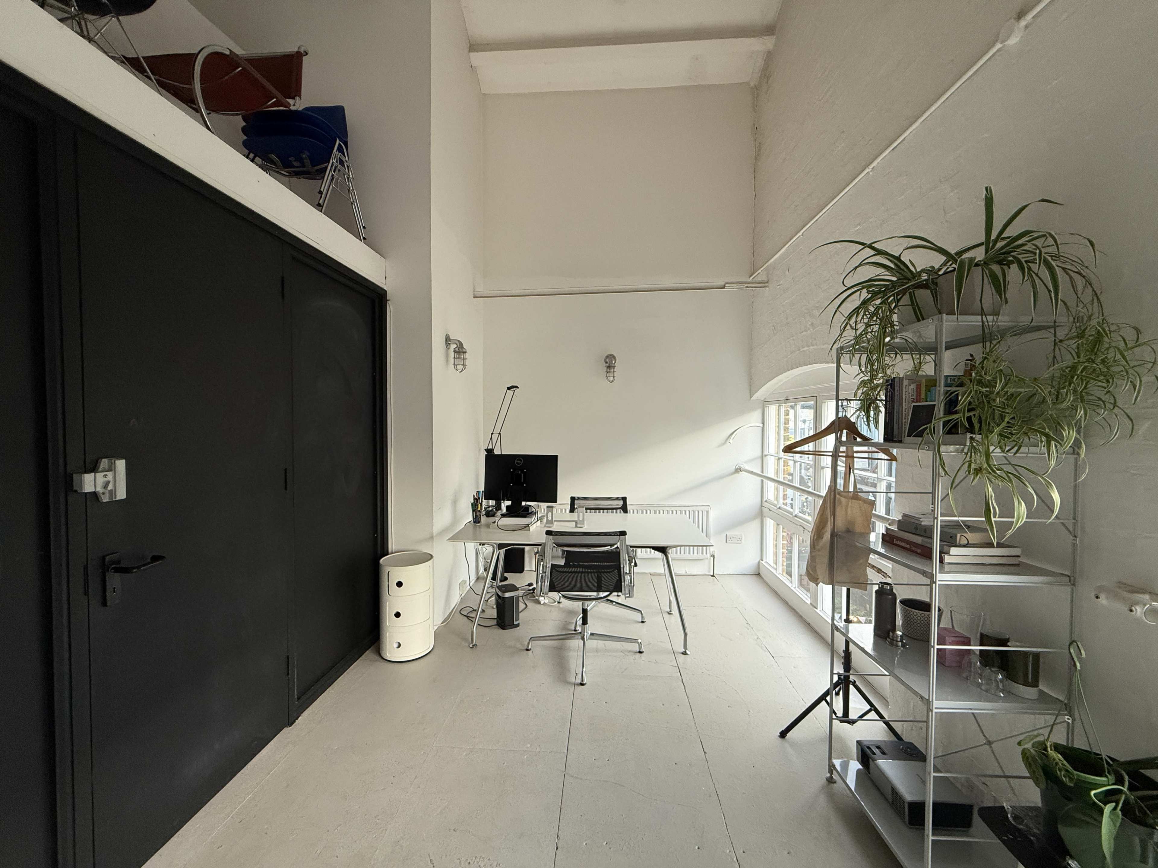 The image shows a modern office space featuring a desk with a computer, a chair, shelving units, and a large window that allows natural light to enter.